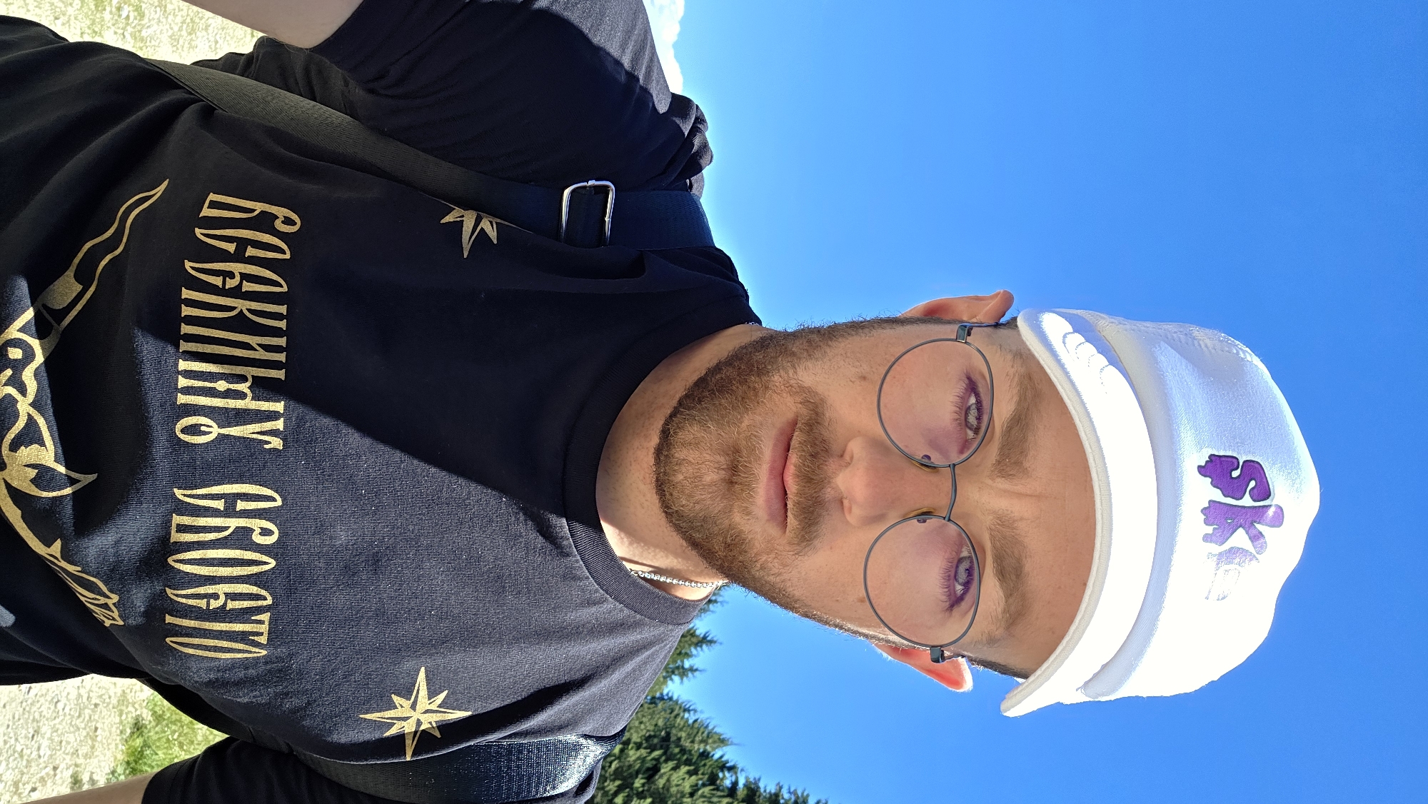 A man wearing a white cap with a purple "SK" logo, round glasses, and a black t-shirt with gold lettering and a star design is captured in a close-up selfie. The background is a bright blue sky with some green trees visible on the right. The man has a beard and a light complexion. He is looking directly at the camera. The lettering on his t-shirt appears to be in a Cyrillic script and is a decorative font.