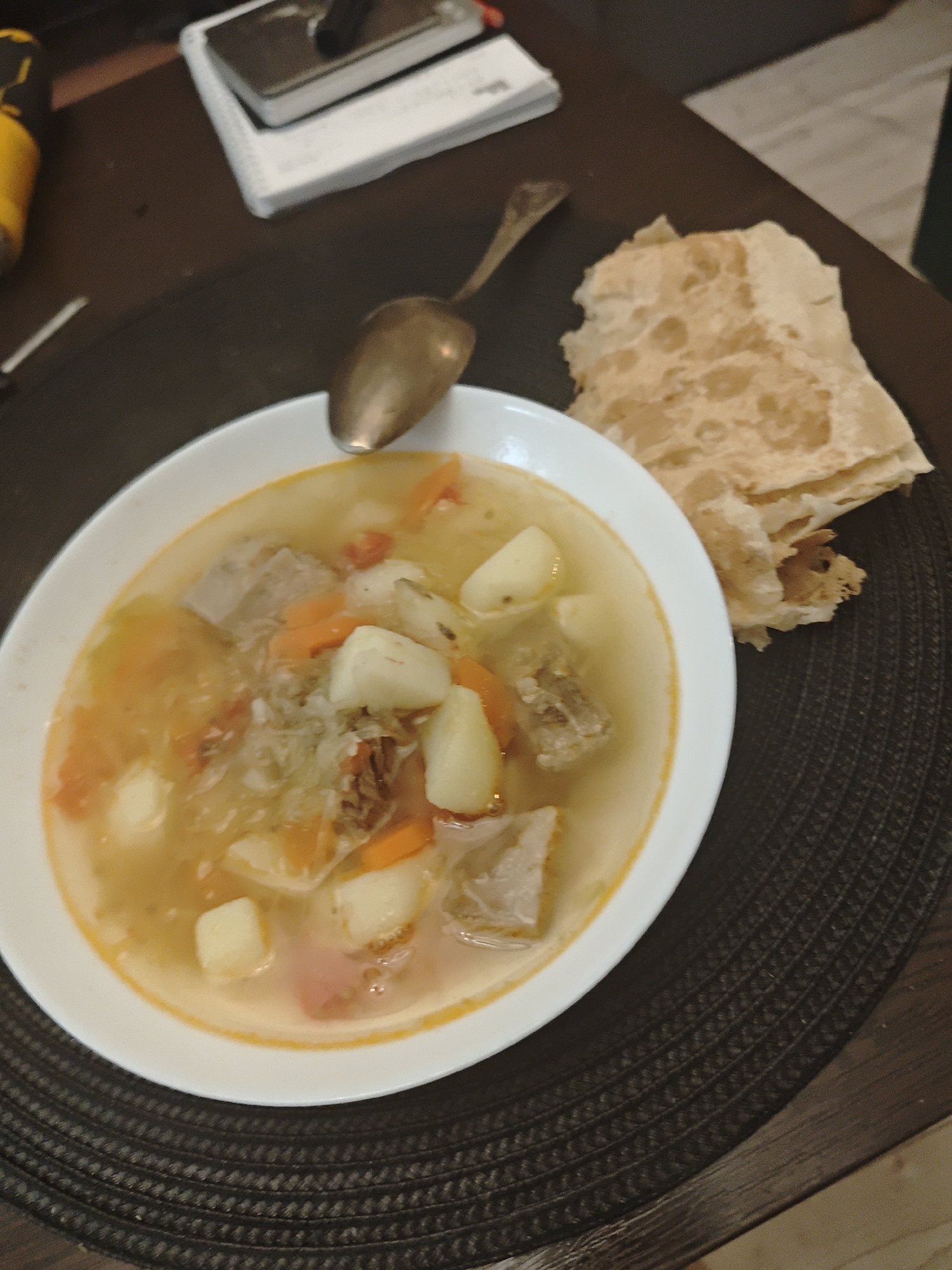 A close-up shot shows a plate of soup, a spoon, and a stack of flatbreads on a dark brown woven placemat. The soup, served in a white bowl, contains chunks of meat, potatoes, carrots, and cabbage in a light broth. An antique spoon rests on the edge of the bowl. A pile of folded flatbreads sits next to the soup. The scene appears to be a meal setting indoors, potentially a home. A notebook and a tool lie out of focus in the background, suggesting a cluttered table. No information indicates the location is in the city of Свистуха (кузяевский С/о), Russia.
