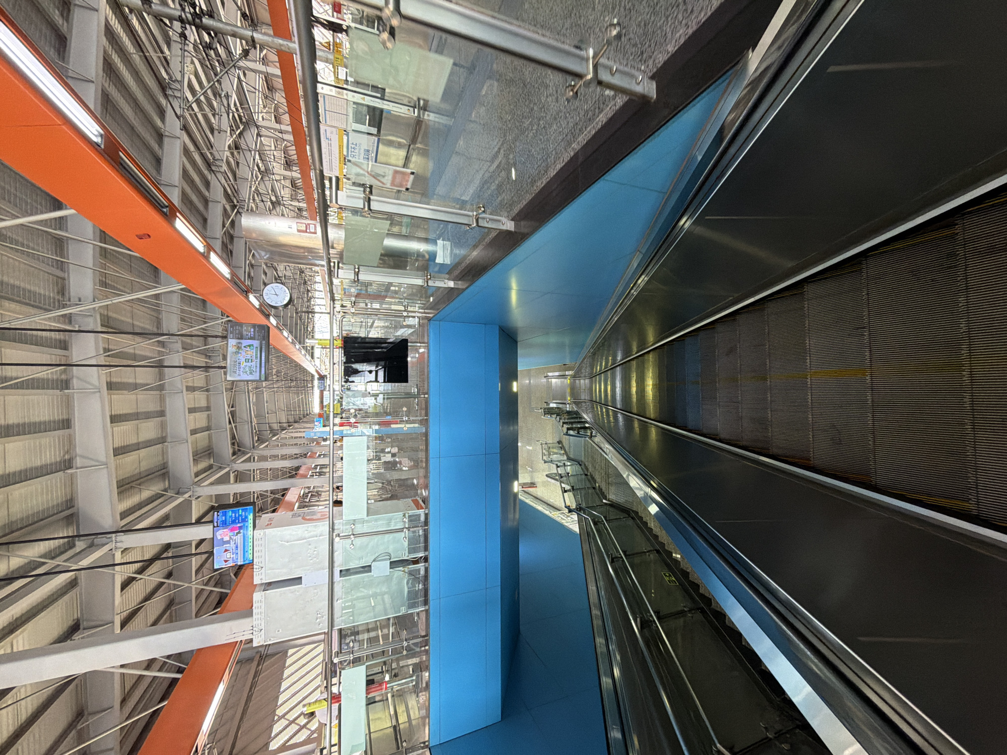 An upward angled shot of a modern subway station escalator in Tiaodeng, China. The escalator is centrally located in the frame, descending into the lower levels of the station. On either side of the escalator, polished metal handrails extend into the distance. The escalator steps are a dark, ribbed metal, with yellow guide lines visible. The surrounding walls of the station are painted a vibrant, almost electric blue. Above, the station ceiling is a complex structure of white beams and orange accents, with industrial lighting fixtures. Glass railings and partitions line the upper platform of the station, reflecting the lights and architecture. A clock is visible on the upper platform, as well as electronic displays showing information. The scene is clean and contemporary, with a focus on functional design. No people are directly visible on the escalator itself.