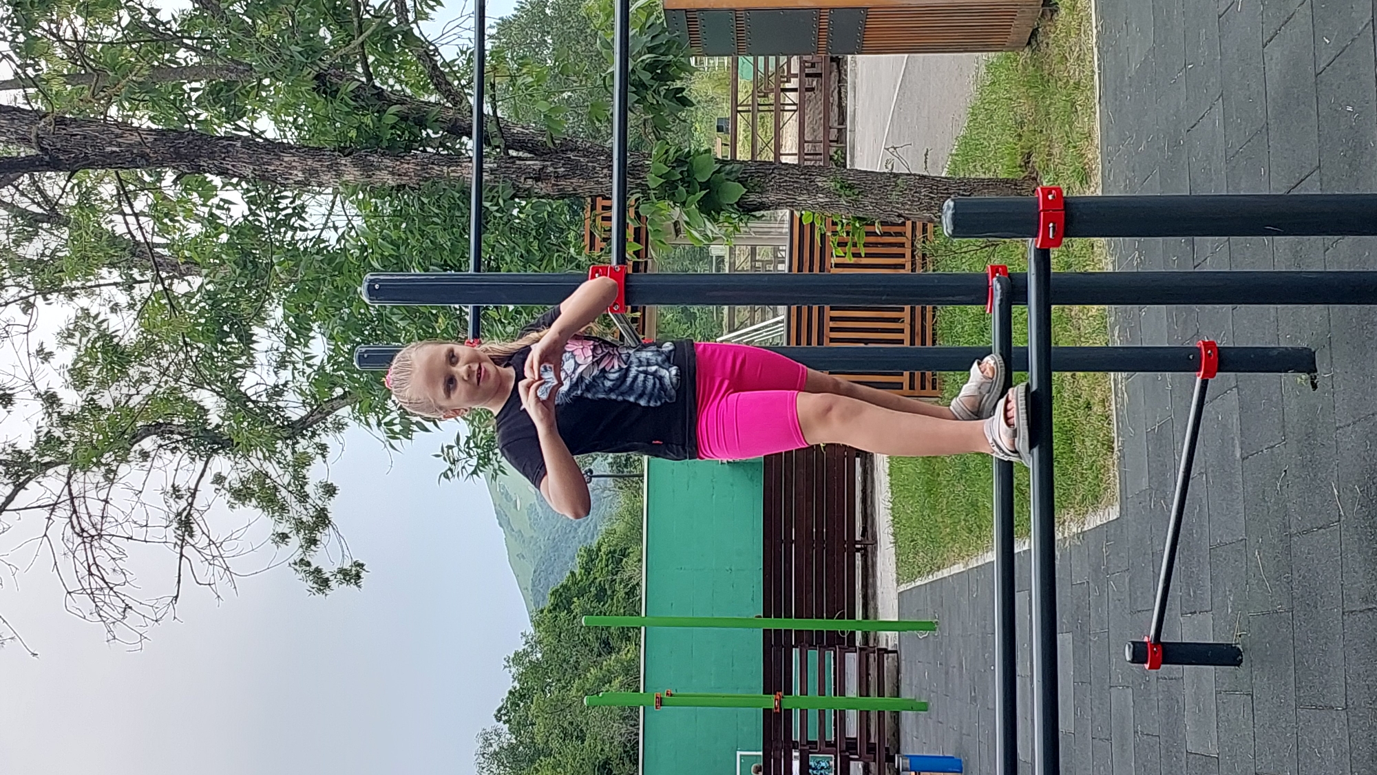 A young girl with blonde hair tied in pigtails is performing a planche on a set of parallel bars in an outdoor gym. She is wearing a black t-shirt with a cat graphic and bright pink shorts. Her feet are resting on the lower bar, and her body is extended horizontally. The gym equipment is made of dark metal bars with red connectors. The background features a large tree with green leaves, a grassy area, and a mountainous landscape under a cloudy sky. In the distance, there are wooden structures and other pieces of gym equipment. The scene appears to be set in the daytime, possibly late morning or afternoon, with overcast weather. The girl's facial expression suggests concentration and effort. The location is identified as Lermontov, Russia, which is known for its mountainous scenery.