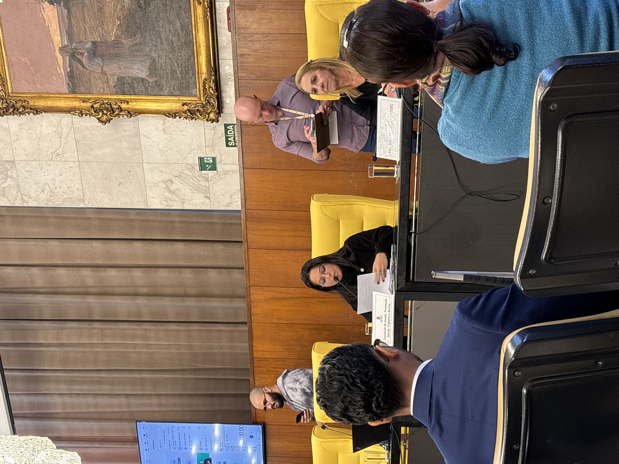 The image captures a meeting setting, likely a parliamentary session or committee meeting, taking place in what appears to be a formal room. Several people are present around a long table. One woman, wearing glasses and dark clothing, is seated and appears to be speaking from a document. A bald man in a purple shirt is standing, possibly presenting or addressing the seated people. Others are seated at the table, some facing the front and listening. There are nameplates with text, "Comissão de Saúde, Promoção Social, Trabalho e Mulher" (Health, Social Promotion, Labor and Women's Committee), suggesting the meeting involves health, social, and labor-related issues. The furniture is yellow. A large painting hangs on the wall behind them, and a 'SAÍDA DE EMERGÊNCIA' (Emergency Exit) sign is visible near the top left of the frame. A monitor screen displays timing information. Based on the signage and Portuguese language text, the location is inferred to be in São Paulo, Brazil.