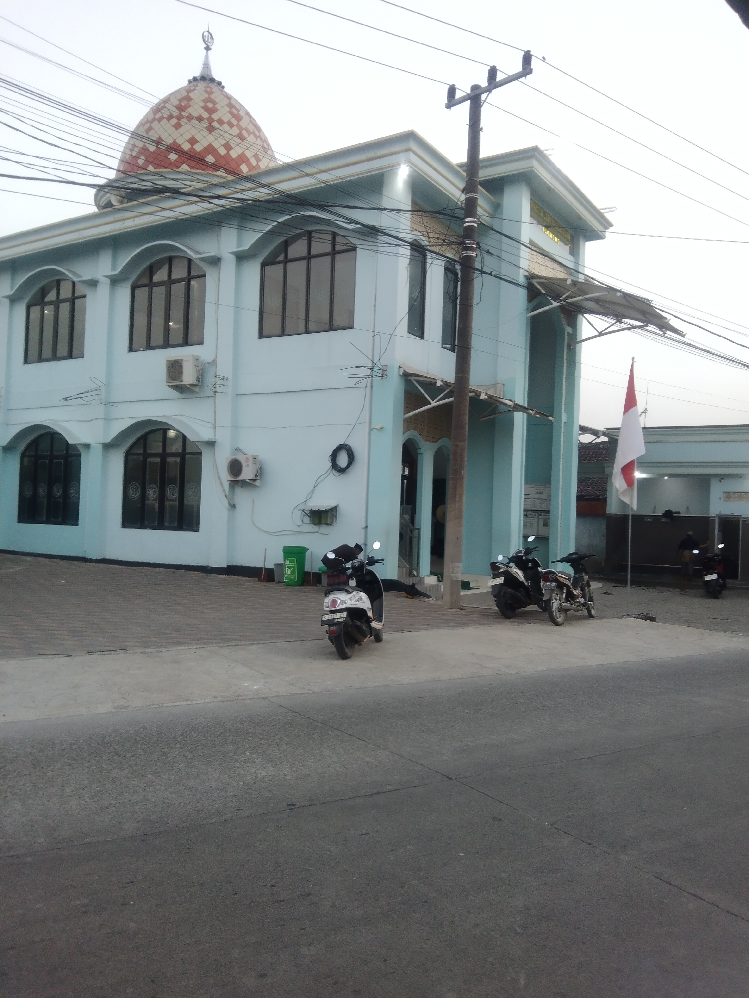 This is a full-shot, eye-level, outdoor, daytime photograph of a light blue building, likely a mosque, in the city of Klepek, Indonesia. The building has a prominent dome, which is red and white checkered, and topped with a finial. There are arched windows on the first two floors of the building, and air conditioning units are mounted on the exterior walls. A utility pole with wires is positioned in front of the building, and several motorcycles are parked in front of it. An Indonesian flag is flying on a flagpole near the entrance of the building. The ground in front of the building is paved with grey tiles, and a road with asphalt is in the foreground. The sky is overcast.