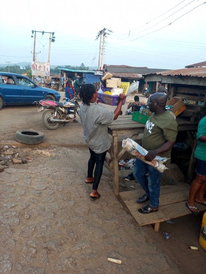 A woman in a grey shirt and black leggings argues with a man in a green shirt holding a bag of rice at an outdoor market. The woman is holding a basket of goods over her head. A blue car and a motorcycle are visible in the background. The market appears to be in a rural area, with buildings and power lines in the distance. An old tire lies on the rocky ground.