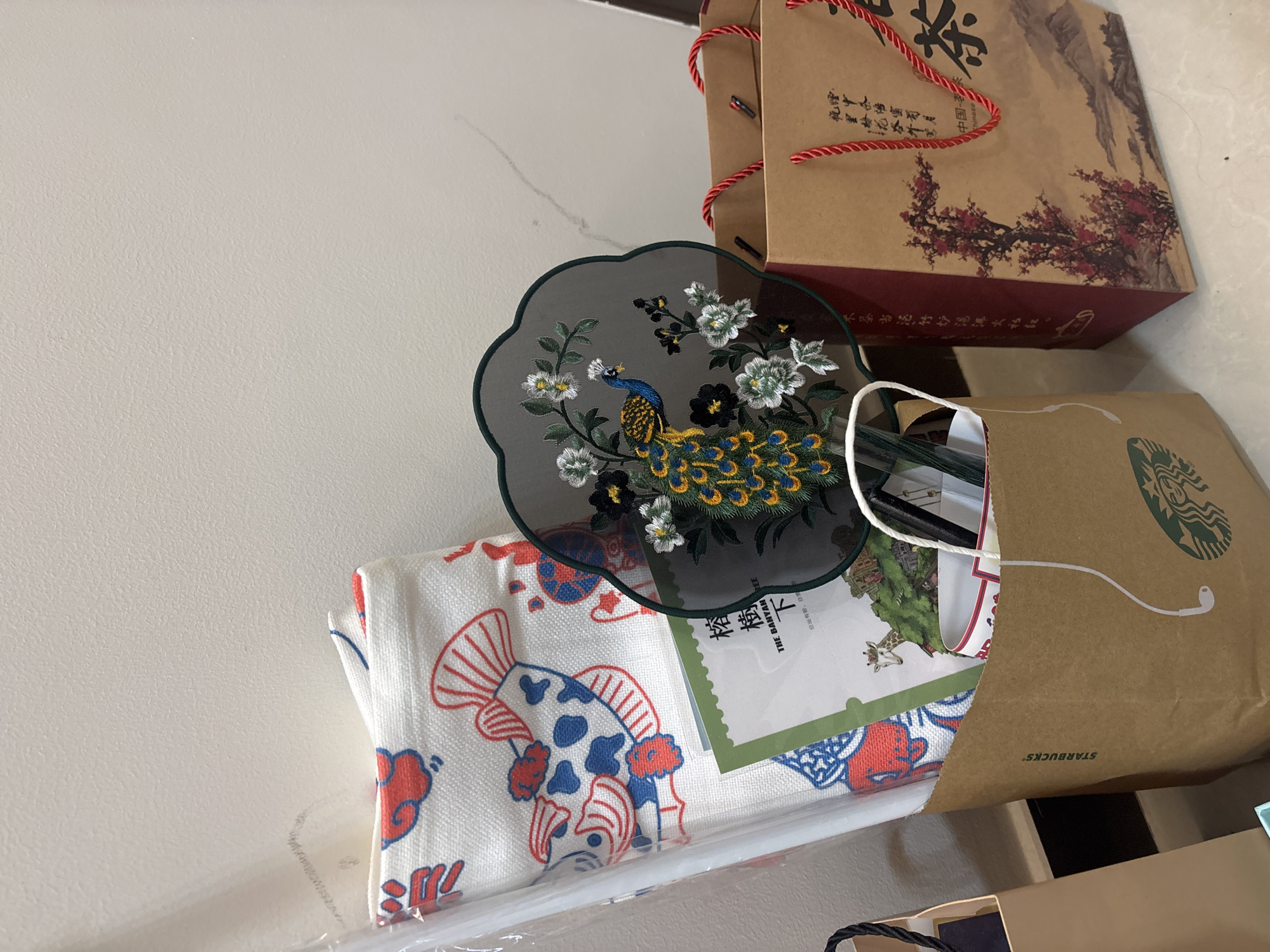 This media file captures a still life arrangement of various items, likely placed on a shelf or table against a light-colored, textured wall. The primary focus is on a collection of bags and decorative objects.
In the background, a tall, rectangular brown paper bag is prominently displayed. It features a red floral design, possibly plum blossoms, and Chinese characters, indicating it might contain tea. A red, twisted rope handle is visible. To its right, a green Starbucks paper bag is partially visible, with its iconic circular logo peeking out.
In the foreground, a decorative fan with intricate embroidery is positioned. The fan is dark in color and features a vibrant depiction of a peacock amidst white and yellow flowers. Below the fan lies a white fabric with a distinctive pattern of red and blue koi fish and other motifs. A green card, also with some Chinese text and an illustration of a giraffe, is tucked beneath the fan and fabric.
The overall scene suggests a curated display of gifts or collected items. The lighting appears to be indoor, with no indication of the time of day or weather. The presence of Starbucks and Chinese characters on the bags points to a setting in China, specifically Tianhe District as per the provided context. There are no people visible, and the arrangement evokes a sense of appreciation for aesthetic and cultural items. The visible text includes "STARBUCKS," "中国," and various Chinese characters on the tea bag and green card.