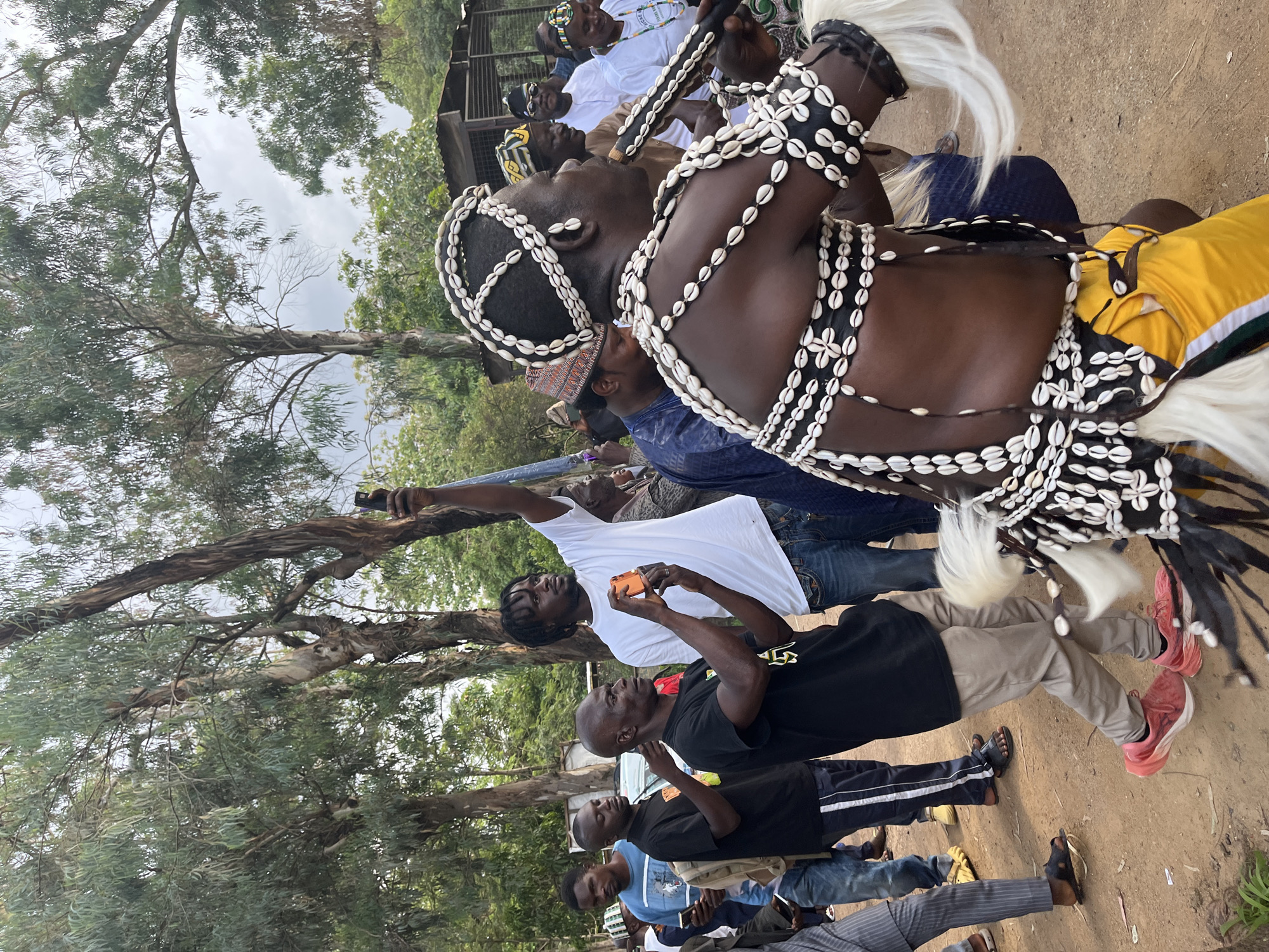 The image depicts a gathering of people outdoors, likely in Jos, Nigeria, under a cloudy sky. In the foreground, a man adorned in traditional attire, featuring elaborate decorations of seashells on his chest, head, and arms, plays a musical instrument that resembles a flute or horn. He wears a yellow and black garment.
Surrounding him are various individuals. Some are actively filming or photographing the performer with their mobile phones. One man in a white t-shirt is holding his phone up, capturing the scene. Another man in a black t-shirt is also taking a picture. Other people are standing and observing the performance.
The environment is characterized by trees with long, slender leaves, suggesting a location with vegetation. In the background, there are some structures that appear to be rudimentary buildings or shelters. The ground is made of dirt. The overall atmosphere seems to be one of cultural observance or celebration.