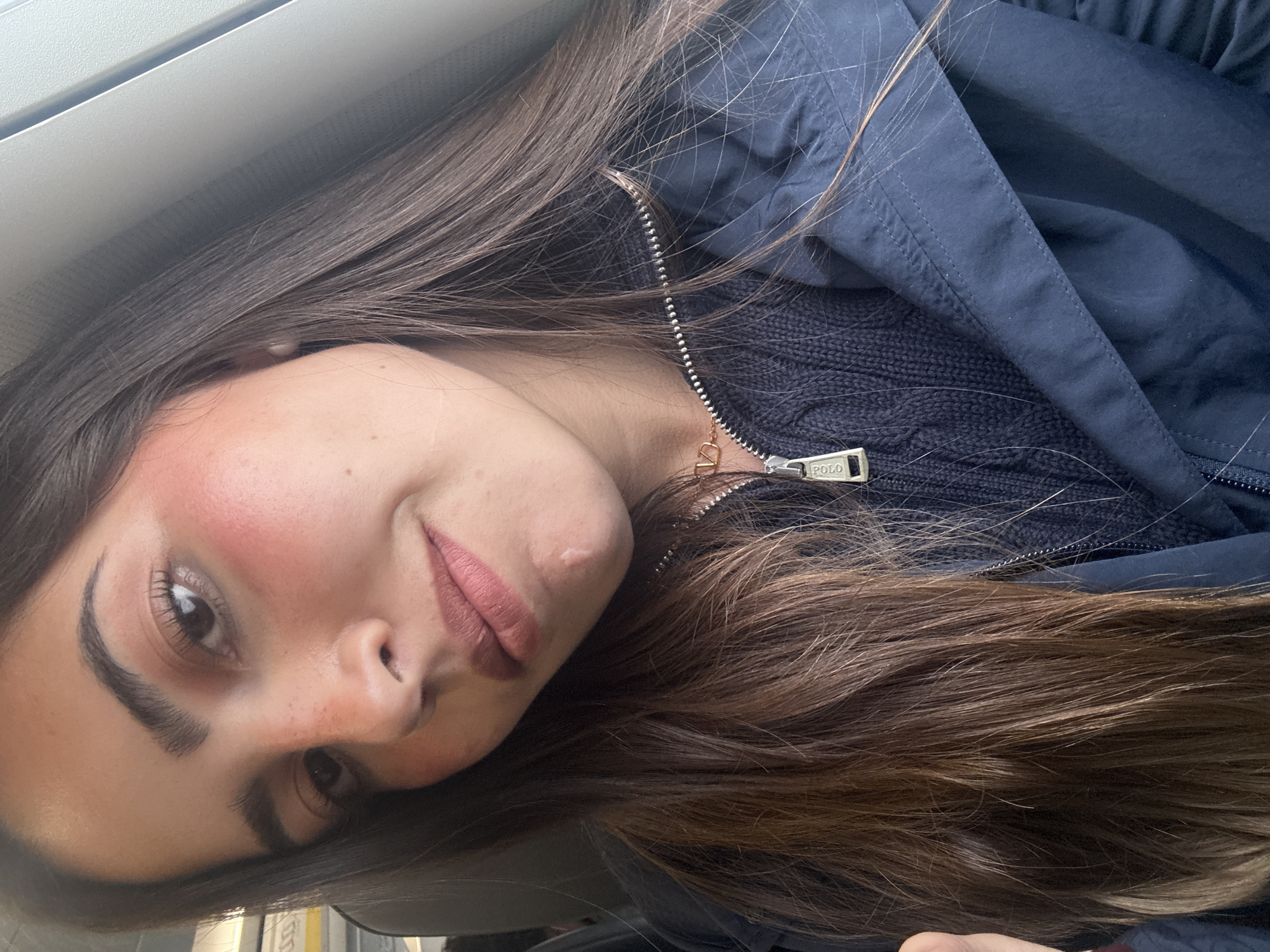 A young woman with long brown hair is pictured from a close-up, slightly angled perspective. She is wearing a dark blue jacket with a visible "POLO" zipper pull. Her makeup includes blush and lip color. The setting suggests she is indoors, possibly near a window, but the exact location within San Antonio, United States of America, cannot be determined from the provided image.