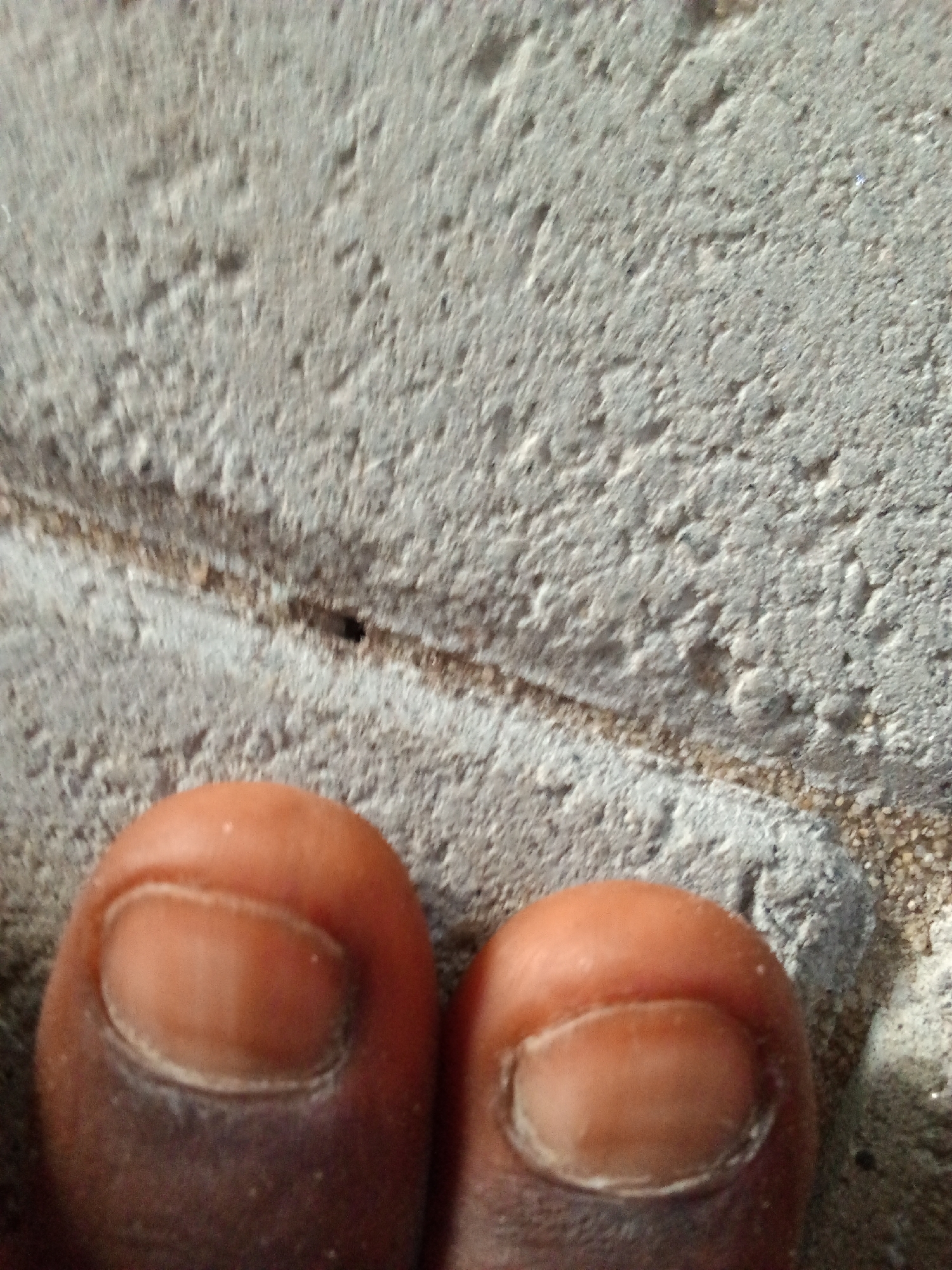 The image shows a close-up view of two human toes resting on a rough, grey, textured surface that appears to be concrete or a similar material. The toes are a darker skin tone. One toe is positioned slightly higher and to the left, with its toenail clearly visible. The other toe is to the right and slightly lower, also showing its toenail. The background is out of focus but consists of the same textured grey surface with some lighter granular particles, possibly sand or small pebbles, visible along a crack or seam in the surface. The lighting suggests an outdoor environment, with shadows indicating a light source from above. The image does not provide any specific landmarks or clues to pinpoint its exact location within Ado, Nigeria, other than it depicts an outdoor ground surface.