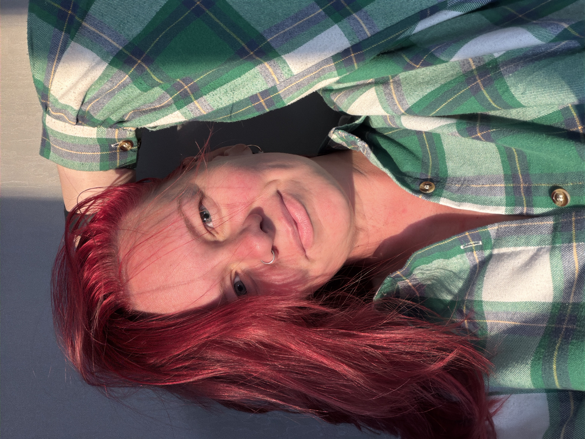A Caucasian female, estimated to be between 25 and 45 years old, is depicted in a medium-close portrait, asserted to be located in the city of Chaikovsky, Russia. However, no specific geographical markers are discernible within the visual frame to corroborate this location. The subject has vibrant, fiery red hair, appearing loose and somewhat windswept, with some strands falling across her forehead and temple. Her eyes are blue, and she gazes directly at the viewer with a subtle, closed-mouth smile. A silver hoop nose ring is visible on her left nostril, and a small silver hoop earring is present in her right ear. Her complexion exhibits some redness on the cheeks, nose, and neck, possibly due to sun exposure or flush. She is wearing a green, blue, white, and yellow plaid flannel shirt, featuring gold-colored buttons on the collar and a cuff, visible on her right arm. Her right arm is raised, with the hand positioned behind or on her head, partially obscured by her hair, appearing to rest against the plain, solid grey background wall. The scene is illuminated by strong, warm, directional light originating from the right side of the frame, creating prominent highlights on her hair and skin, and casting soft shadows across her face and body.