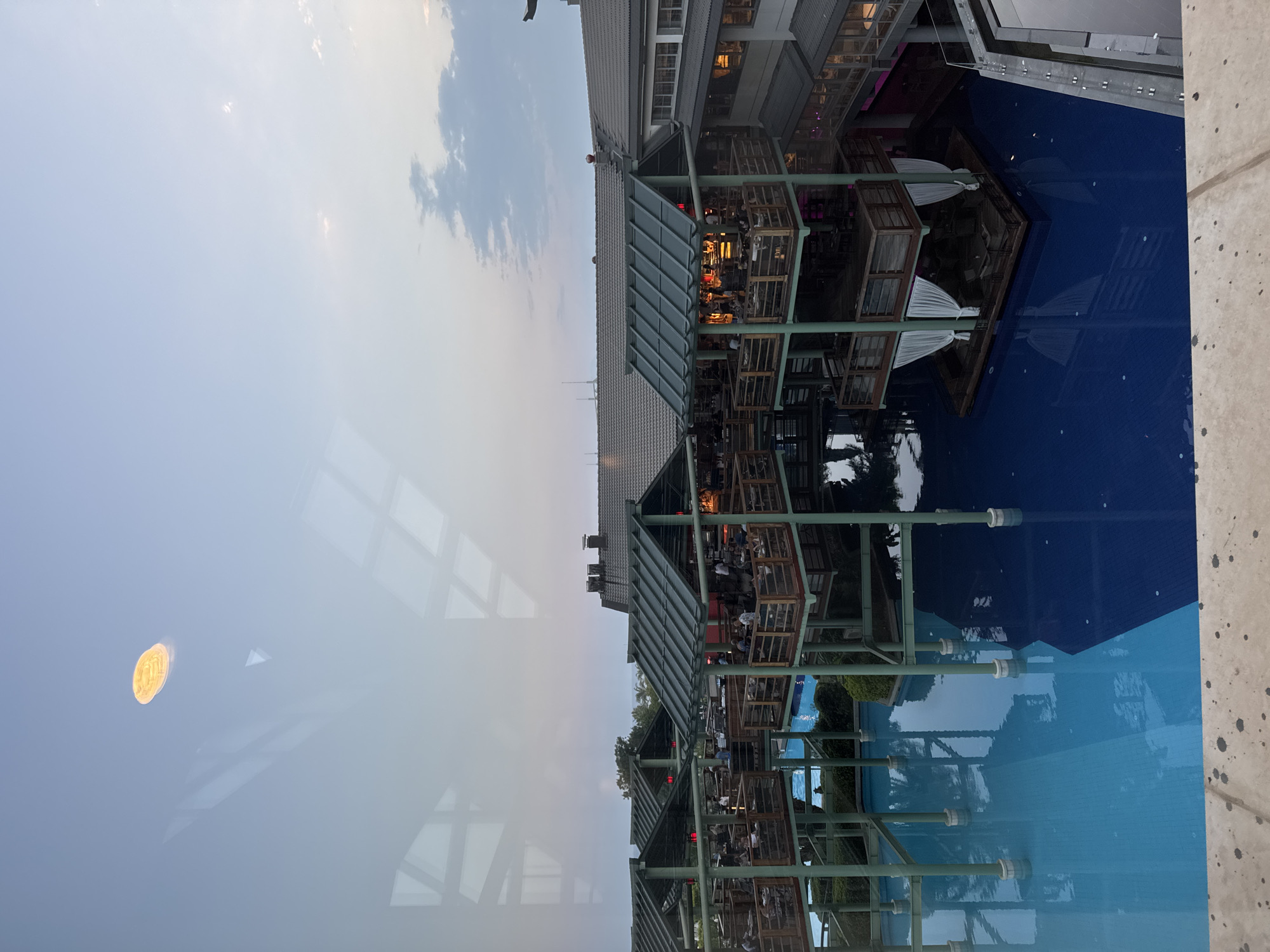 The image shows an elevated, multi-tiered dining structure with a dark, sloped roof, overlooking a deep blue swimming pool. The structure has open-air seating areas with wooden railings, where numerous people are dining. White curtains hang from the lower level of the structure, which also appears to be a seating area. The pool reflects the structure and the sky. In the upper portion of the image, there is a light-colored circular object in the sky, possibly a light or the moon, and reflections from a window are visible. The background shows a building with a dark tiled roof and distant trees. The scene is set during dusk, with a soft, diffused light. The location is identified as Aksu, Turkey.