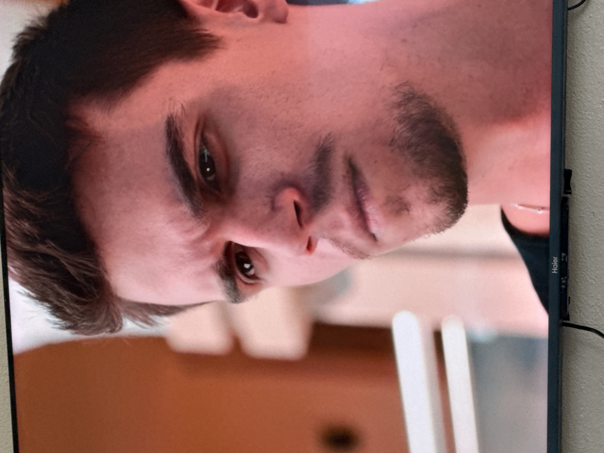The image shows a close-up of a young man's face. He has dark hair, dark eyebrows, and a faint mustache and beard. His eyes are looking to his left, and his expression appears serious or contemplative. The lighting suggests an indoor setting with warm tones. The background is blurred, but appears to be some sort of structure with light-colored elements. The bottom of a television screen with the "Haier" logo is visible at the very bottom of the image, indicating the man's face is likely a still from a video being displayed on the screen. There is no discernible information about the city of Uyutnoye, Ukraine, within the image itself.