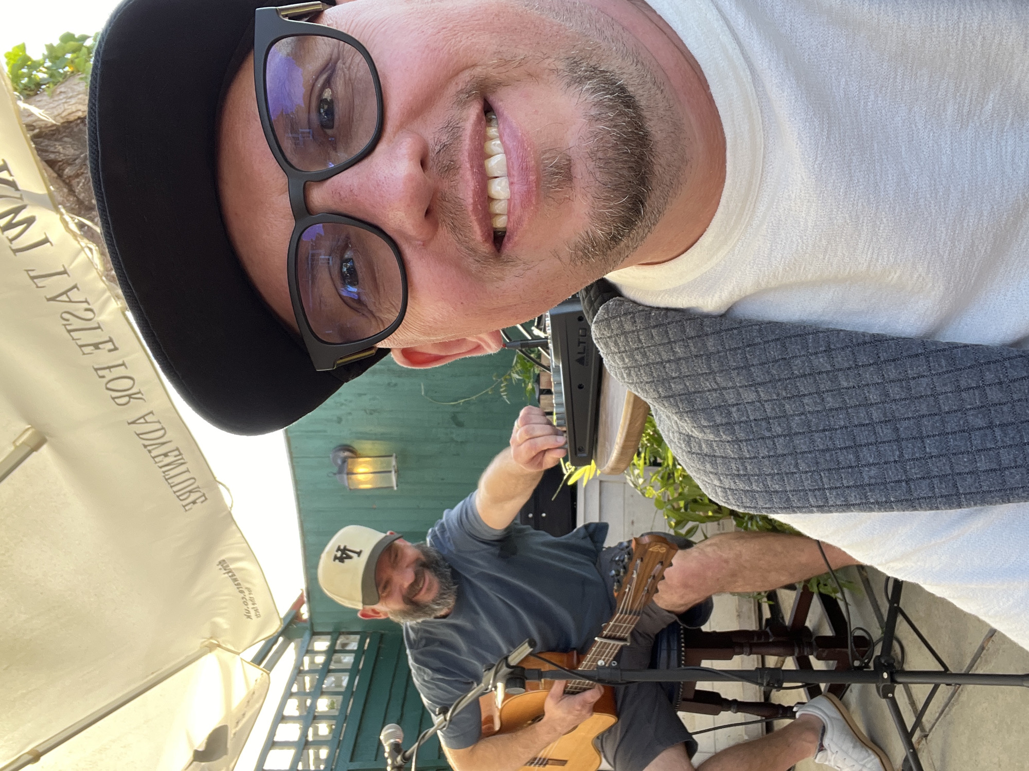 A man in a white t-shirt and grey cardigan, wearing glasses and a black baseball cap, is taking a selfie outdoors, with another man in the background playing a guitar. The second man is wearing a blue t-shirt and a beige baseball cap with the letters "LA" on it. He is seated on a stool, holding an acoustic guitar and interacting with a sound mixer on a table. The environment appears to be an outdoor seating area, possibly a pub or restaurant, with green wooden fencing and a white umbrella overhead. The umbrella has black text that reads "TASTE FOR ADVENTURE". The image is taken in the city of Guildford, United Kingdom.