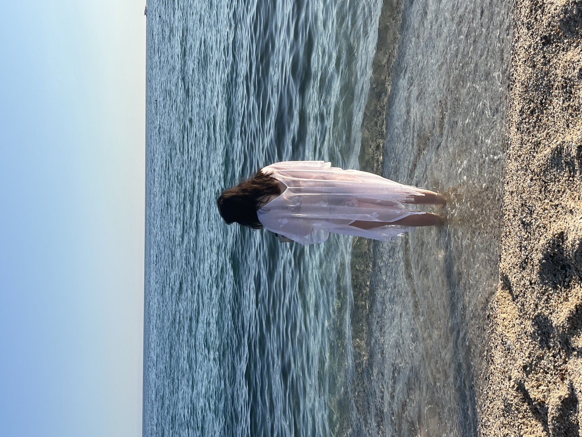 The image shows a woman standing in the water at a beach in Alanya, Turkey. The water is clear and blue, and the sky is a light blue. The beach is made up of small pebbles. The woman is wearing a white, sheer dress. She has dark hair. She is standing with her back to the camera. The image is taken from a slightly elevated perspective.