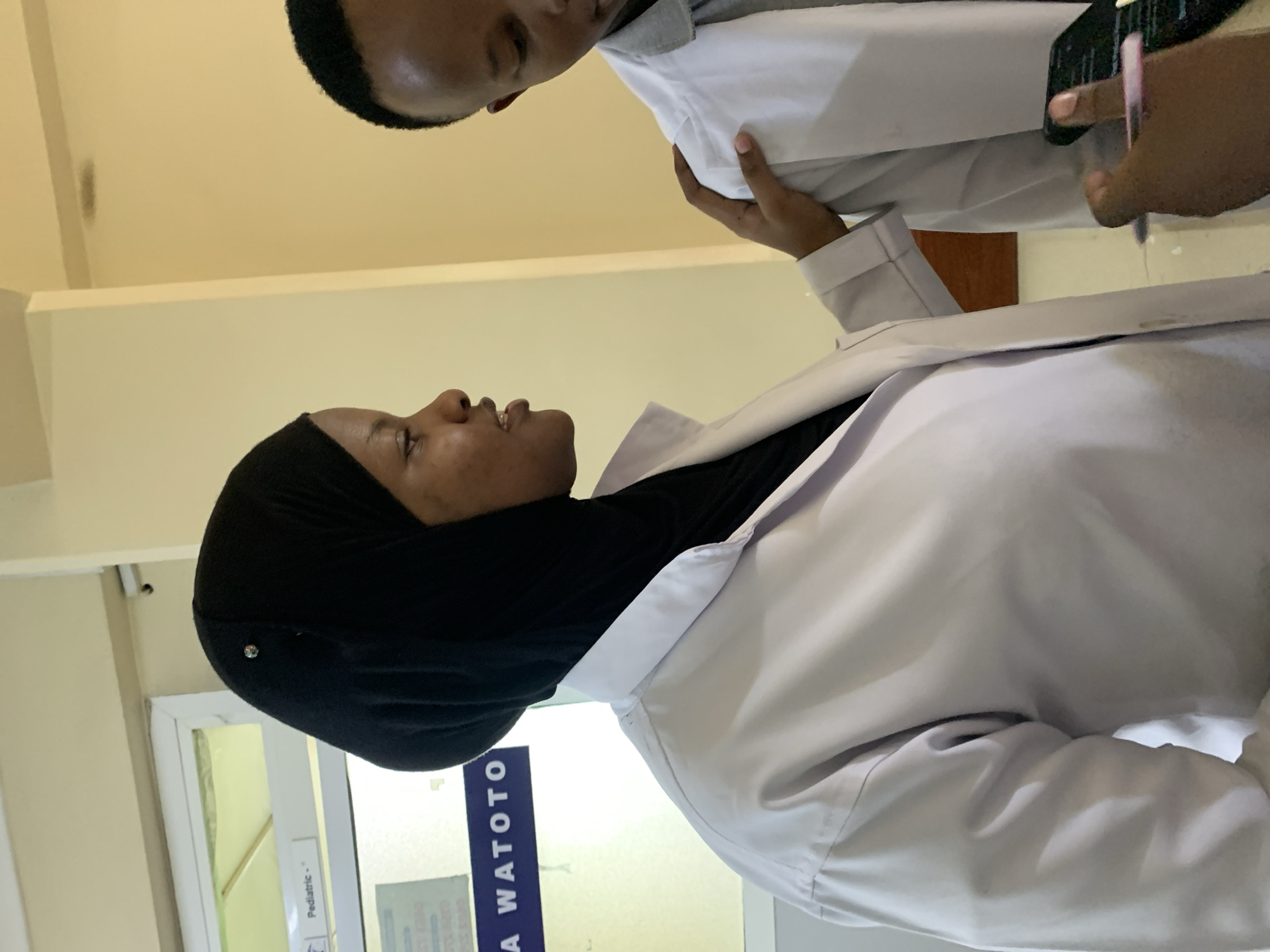 Two people are in a medical setting. The person closest to the camera is wearing a black headscarf and a white coat. The other person is wearing a white coat and is holding a mobile phone. The location appears to be the Pediatric section of a clinic or hospital labeled in Swahili with the word "WATOTO", suggesting it is in Tanzania, most likely Dar es-Salaam.