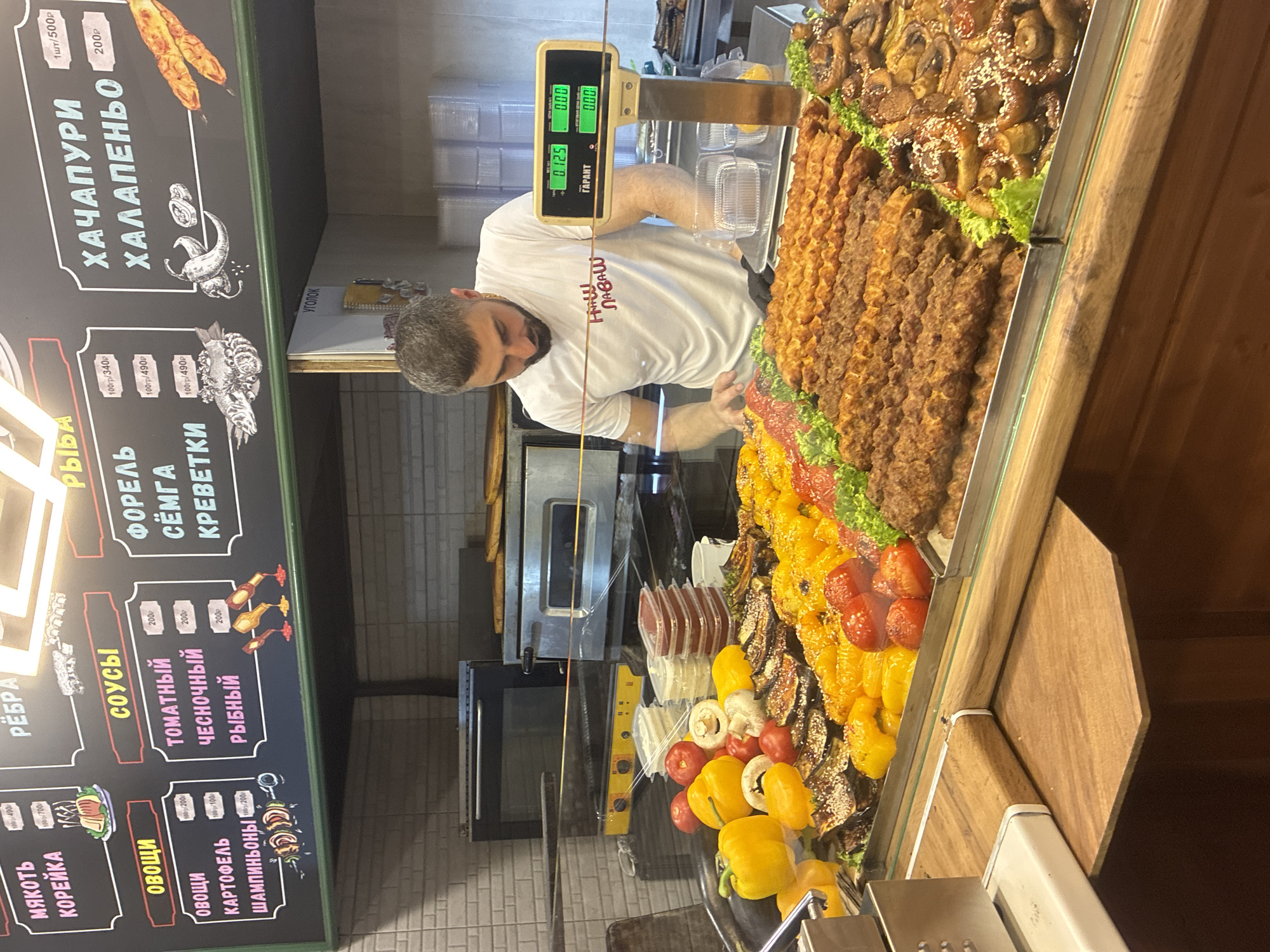 In Sirius, Russia, a male employee in a white t-shirt with red lettering stands behind a refrigerated display case filled with various food items. The display case contains rows of grilled meats on skewers, grilled vegetables such as bell peppers and eggplant, and what appears to be grilled mushrooms. The employee is focused on the scale in front of him, which displays digital readings. Behind the employee, a menu board is visible, featuring sections with text in Russian, including "МЯКОТЬ КОРЕЙКА", "ОВОЩИ", "СОУСЫ", "РЫБА", and "ХАЧАПУРИ ХАЛАПЕНЬО". The environment suggests a food service establishment, possibly a restaurant or a deli, with kitchen equipment such as ovens visible in the background. The overall scene depicts a busy food preparation and service area.