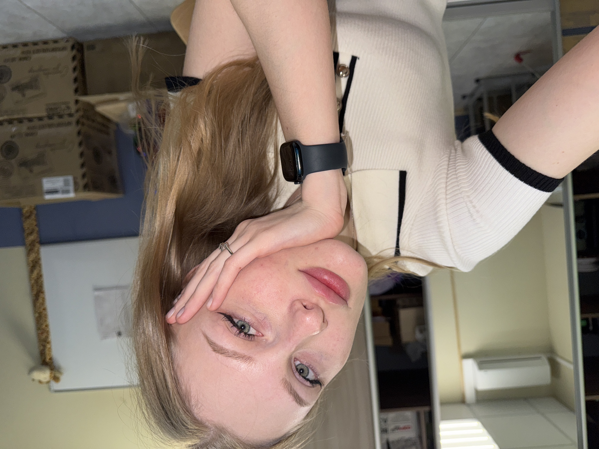 A fair-skinned young woman with blond hair and green eyes is seen close-up, with her head tilted to the right. She is wearing a white knit top with black trim and an Apple watch on her left wrist. Her left hand is resting on her forehead, and she is wearing a ring on her ring finger.
In the background, partially visible, are cardboard boxes stacked in the upper left corner. There is also a white board and a scratching post. A reflection suggests a room interior with an air conditioning unit on the wall. The city of Perm, Russia, is not indicated in the image.