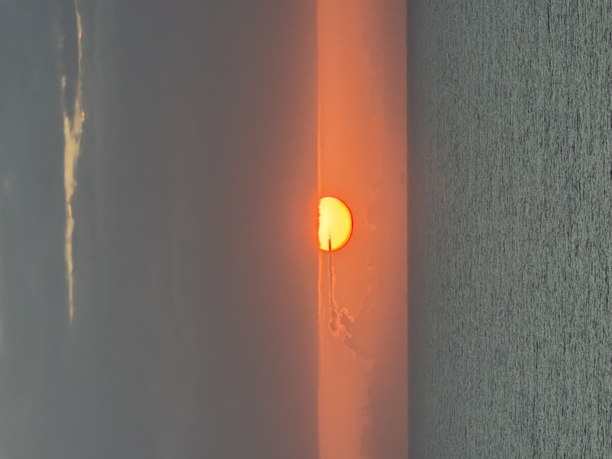 The image captures a vibrant sunset or sunrise over a tranquil body of water, likely an ocean or a large lake. The scene is bisected horizontally into the sky and the water.
In the upper portion, a large, intensely glowing sun, appearing in bright orange and yellow, is partially obscured by clouds or atmospheric haze as it dips towards or rises from the horizon. The sky immediately surrounding the sun is awash in rich hues of orange and red, indicating the early or late hour. Above this fiery glow, the sky transitions to softer greys and blues, with wispy, elongated clouds visible, some catching the light with lighter streaks.
The lower half of the image presents the surface of the water, characterized by a textured appearance suggesting subtle ripples or small waves across its expanse. The water reflects the ambient light, taking on muted tones of grey and blue. The overall impression is one of serene beauty, with the hazy atmosphere contributing to the diffused light and soft coloration of the sky.
There are no people or specific objects discernible, and no visible text. The time of day is clearly either dawn or dusk, and the weather appears hazy or slightly cloudy, enhancing the sun's ethereal glow.