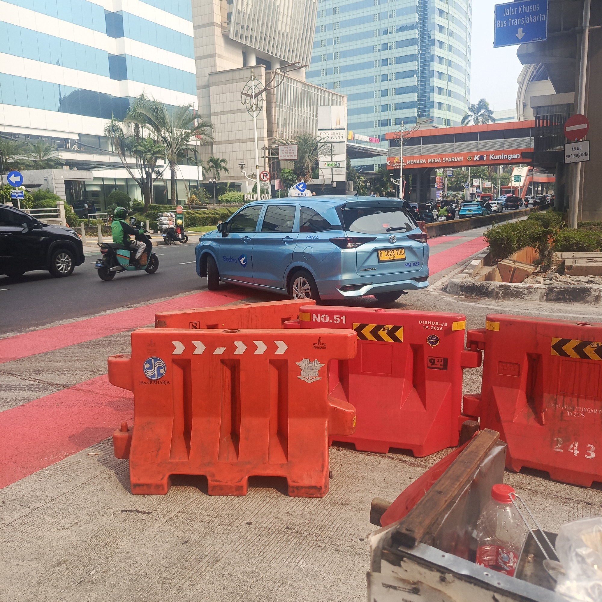 The image depicts a street scene in South Jakarta, Indonesia, likely in the vicinity of Kuningan, given the signage visible. The foreground is dominated by bright orange traffic barriers. Behind the barriers, a blue taxi, identified as a Bluebird, is driving on a road marked with red lines. A person wearing a green jacket and helmet is riding a scooter in the same direction as the taxi. In the background, modern high-rise buildings with glass facades are visible, along with palm trees and other urban vegetation. A pedestrian bridge with signage indicating "MENARA MEGA SYARIAH" and "KUNINGAN" spans over the road. A blue street sign indicates "Jalur Khusus Bus Transjakarta" (Transjakarta Bus Special Lane) with a downward arrow. The overall impression is a busy urban environment with ongoing traffic and construction or traffic management activities.