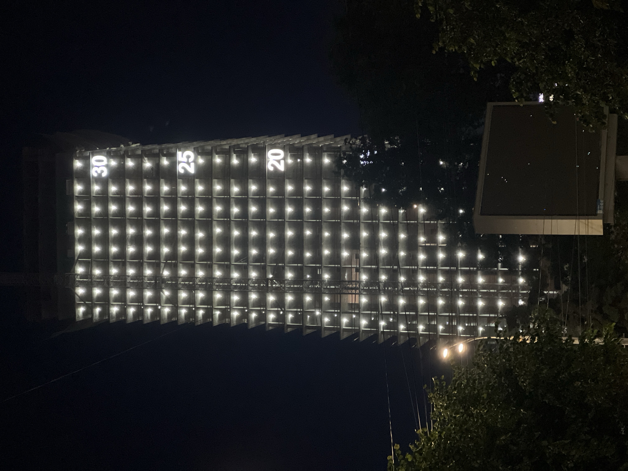 The image displays a large, modern building brightly illuminated at night, extending horizontally across the frame. Its facade is covered by a dense grid of numerous square-shaped, glowing white lights, creating a striking and patterned illumination against the dark sky.
Prominently featured on the upper part of the building are large, vertically oriented, glowing white numbers: "30", "25", and "20". These numbers appear to be part of the building's design, possibly indicating floors or a countdown.
To the right side of the illuminated structure, a dark, rectangular billboard or large screen is visible, partially obscured by dense, dark foliage from trees. More tree branches and leaves, along with several thin wires or cables, are visible in the foreground, particularly on the right side of the image.
The scene is clearly set at night, with the strong artificial lighting of the building casting a stark contrast against the dark background. No people or specific activities are discernible. The overall impression is one of a modern urban structure, possibly in Izhevsk, Russia, as indicated by the location context.