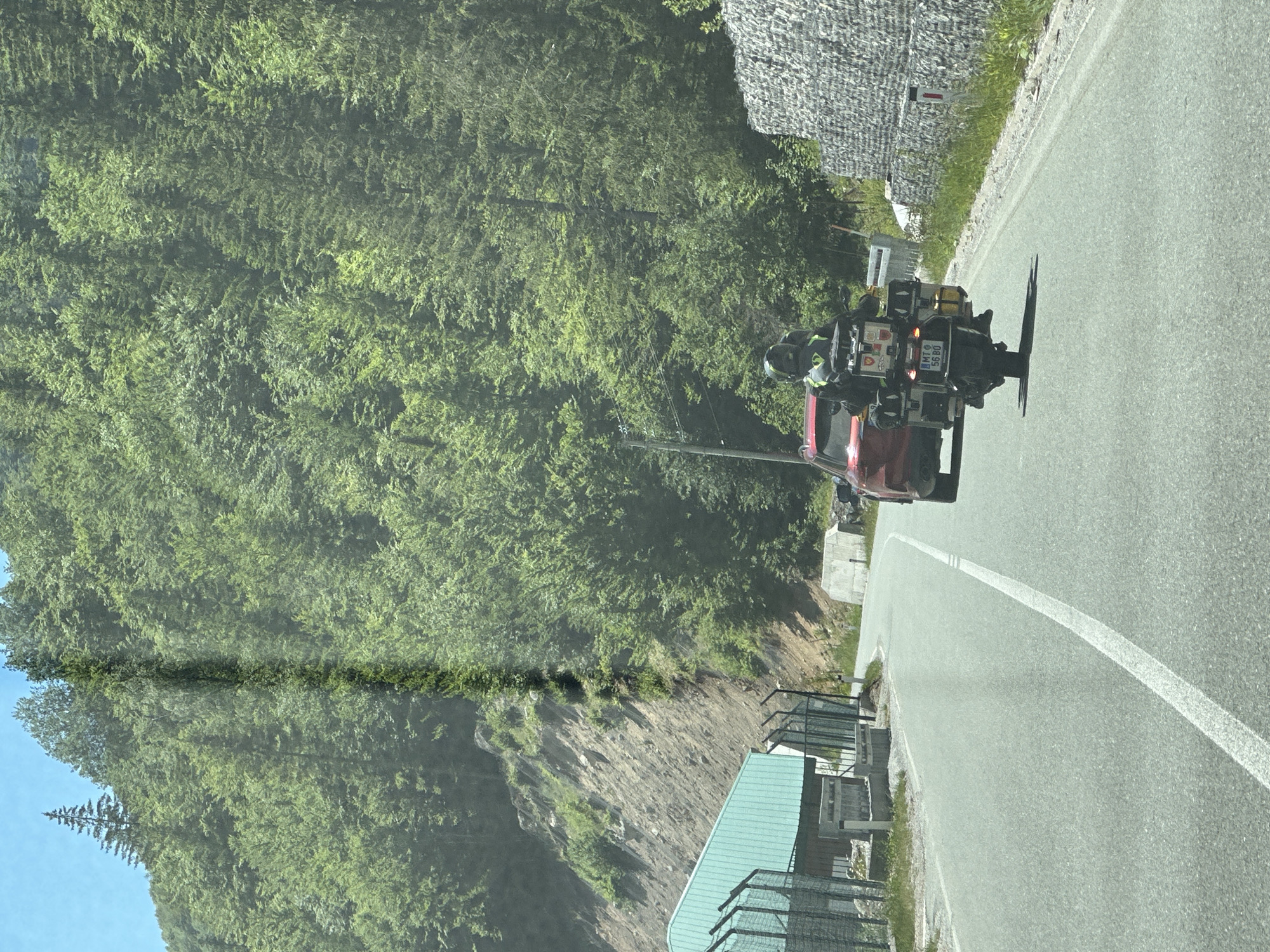 In the city of Lubnice, Montenegro, a red SUV is driving on a paved road lined with a forest of green trees on a steep hillside. A motorcycle with a rider wearing a helmet and a yellow vest is behind the SUV. The motorcycle is equipped with multiple storage boxes and a license plate that reads "MTO 56 80". On the left side of the road, there is a light blue building with a fence in front of it. The sky is clear and blue.