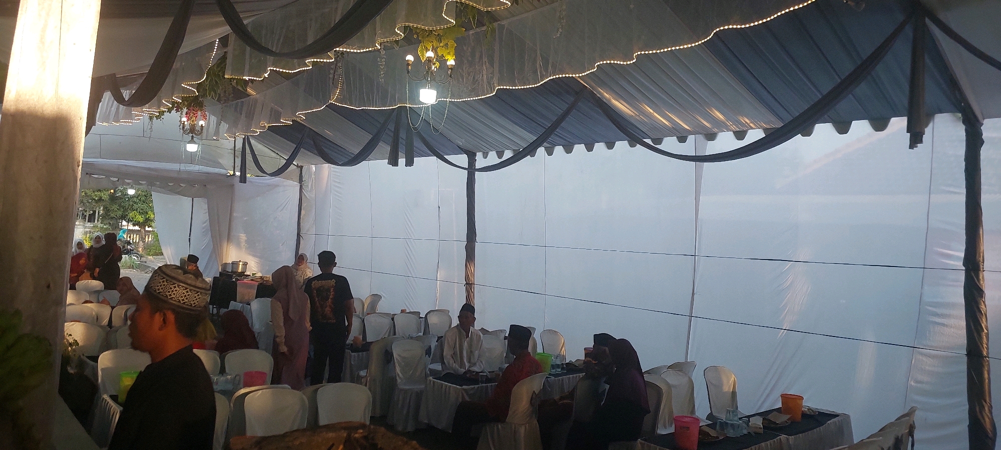 The image shows an outdoor event space, possibly a wedding reception or a similar gathering, in Bukur, Indonesia. The scene is under a large tent with white and dark blue fabric draped across the ceiling. String lights and a chandelier with hanging plants illuminate the space. Several people are seated at tables covered with black tablecloths. Some individuals are standing and interacting. The environment appears to be a partially enclosed area, with white fabric walls on the right side of the frame and an open area with trees and a building visible on the left. The lighting suggests it is either late afternoon or early evening.