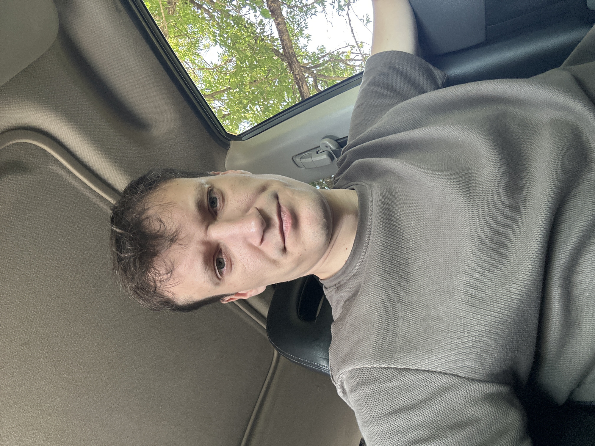 A man with short, dark, messy hair and a light complexion is seated in the driver's seat of a vehicle. He is looking directly at the camera with a neutral expression. He is wearing a dark grey, short-sleeved t-shirt with a textured pattern. The interior of the car is visible, including the dark grey headliner and a black leather headrest of the passenger seat. The window on the right side of the vehicle is open, revealing green foliage and tree branches against a bright sky, suggesting an outdoor, possibly rural setting. The time of day appears to be daytime. The specific location in the city of Makiv, Ukraine, cannot be determined from the visual information present in the image.