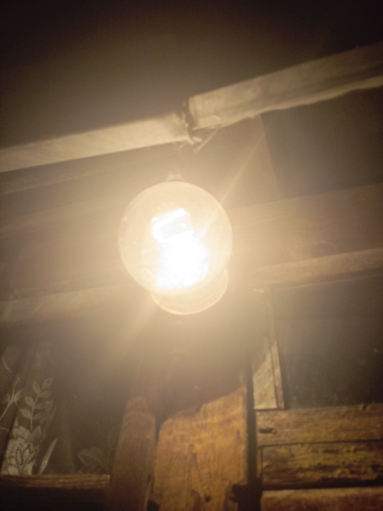 A dimly lit interior scene features a clear glass light bulb suspended from a wire, emitting a bright, warm light that illuminates the surrounding wooden beams and structures. The background is dark, with hints of patterned fabric on the left side. The overall atmosphere is rustic and somewhat aged. The location is identified as Ухманы, Russia. There are no people or animals visible in the frame.