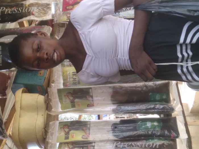 This image depicts a young Black female standing within a retail stall in Agege, Nigeria. The female subject, positioned slightly right of center, wears a white, ruched, short-sleeved crop top and a black skirt with white horizontal stripes near the hem. Her dark hair is short. Her mouth is slightly open, suggesting she may be speaking or in mid-sentence. Her left hand is visible, holding a black plastic bag.
The background reveals the commercial nature of the scene. To the left and behind the subject, multiple long, clear plastic packages of hair extensions are hanging vertically. These packages prominently display "X-Pression" and "RICH BRAID" branding, containing hair in various shades, including black and brown, with small green labels visible within the packaging. Above these, an overturned, light-colored plastic basin rests on a shelf. Behind the basin, stacked blue and brown cardboard boxes are present. To the right of the subject, more packaged hair extensions, some with "LUSCIOUS" branding, are hanging. The environment suggests an outdoor or semi-enclosed market stall, with daylight illuminating the scene.