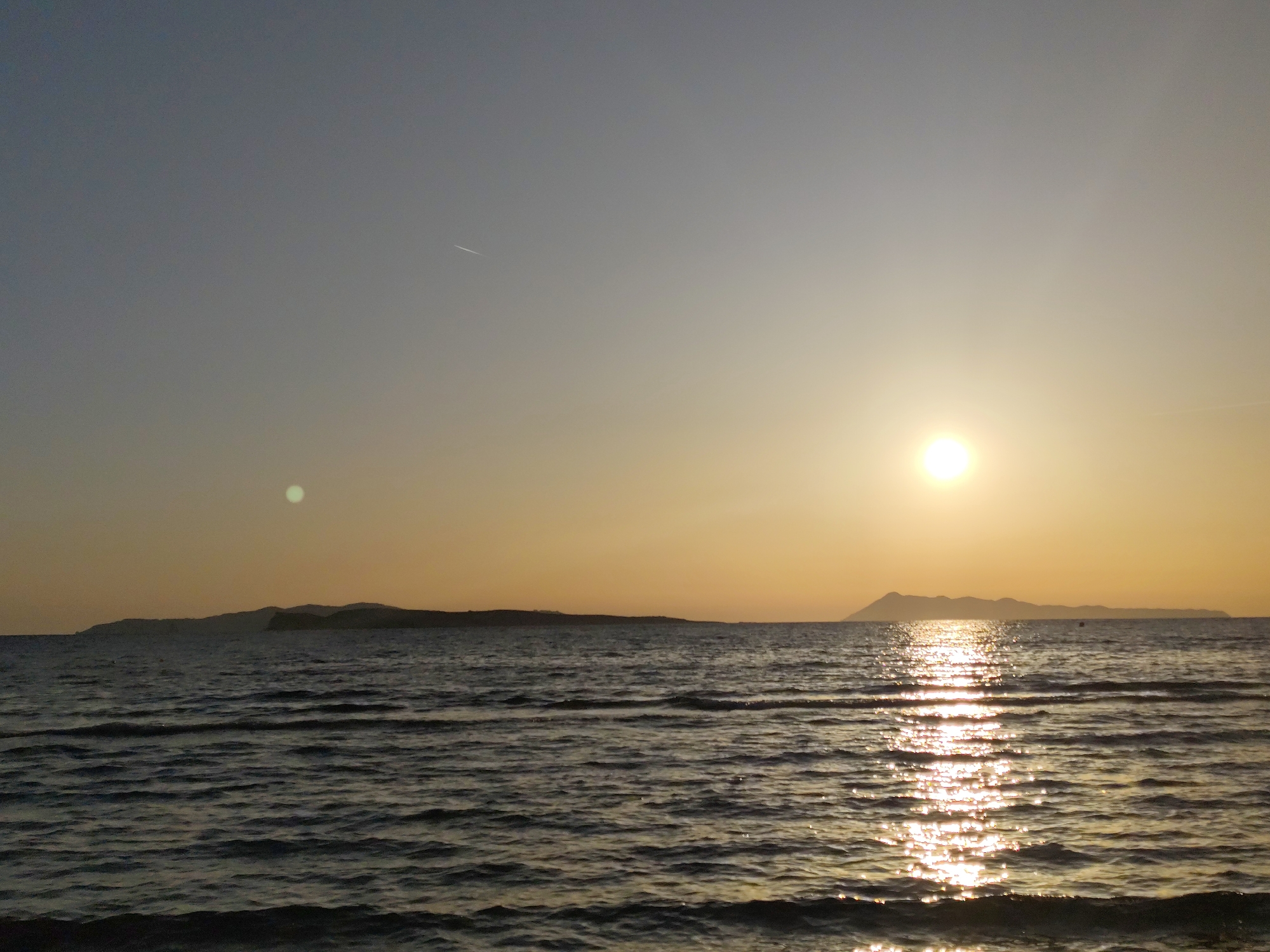 The image depicts a sunset over the sea, in an unknown location of an unknown city. The sun is setting on the right side of the image, casting a bright glare on the water. There are two islands visible in the distance. The sky is a gradient of colors, ranging from light blue to orange near the horizon. The water is dark and choppy, with some reflections of the sun. There is a visible jet stream from an airplane in the sky above.