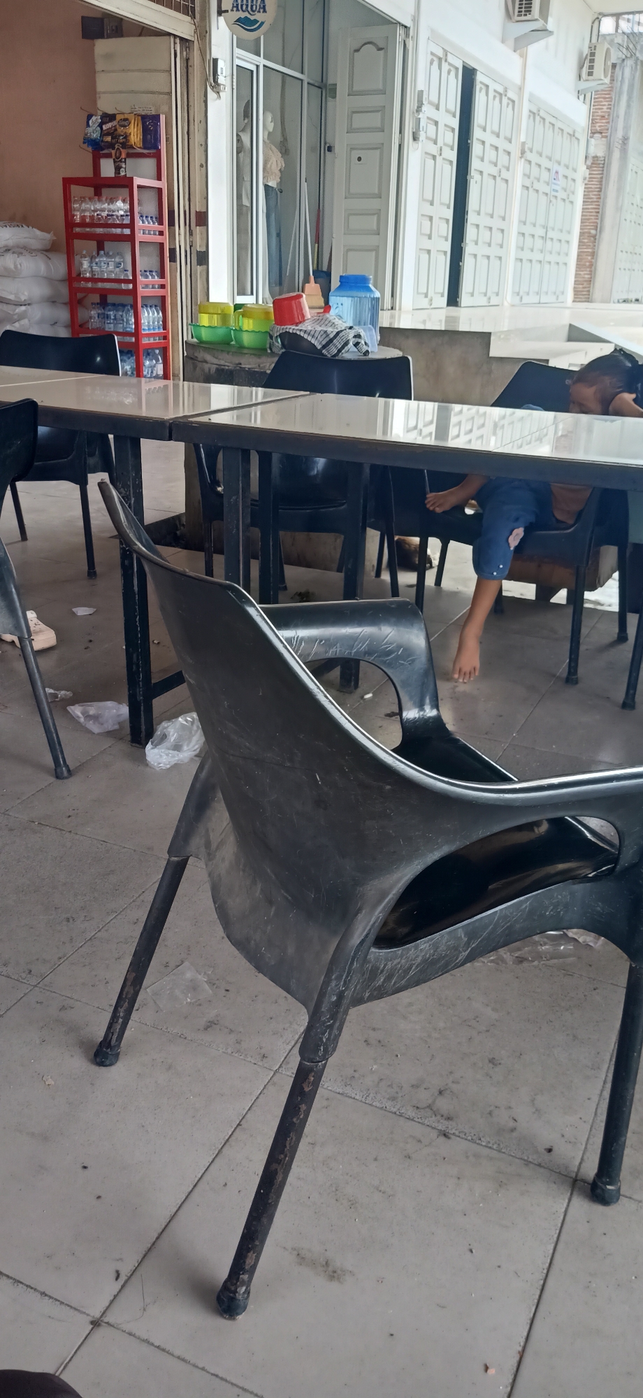 In Tambonbaroh, Indonesia, an outdoor seating area is depicted, likely associated with a shop or eatery. The foreground is dominated by a black plastic chair, tilted forward, with its metallic legs showing signs of wear. Beyond it, a long, dark table with a reflective surface is visible, supported by dark metal frames. Several other black plastic chairs are scattered around the table, some occupied. In the background, a store interior is visible through glass doors, featuring a red shelving unit stocked with bottles and packets, and mannequins dressed in clothing. To the right, a young person is seated at the table, their legs visible beneath it, resting their head on their arms. The ground is tiled and appears somewhat dirty, with scattered plastic bags. A blue water dispenser and some colorful plastic containers are present on a raised platform to the left of the store entrance.