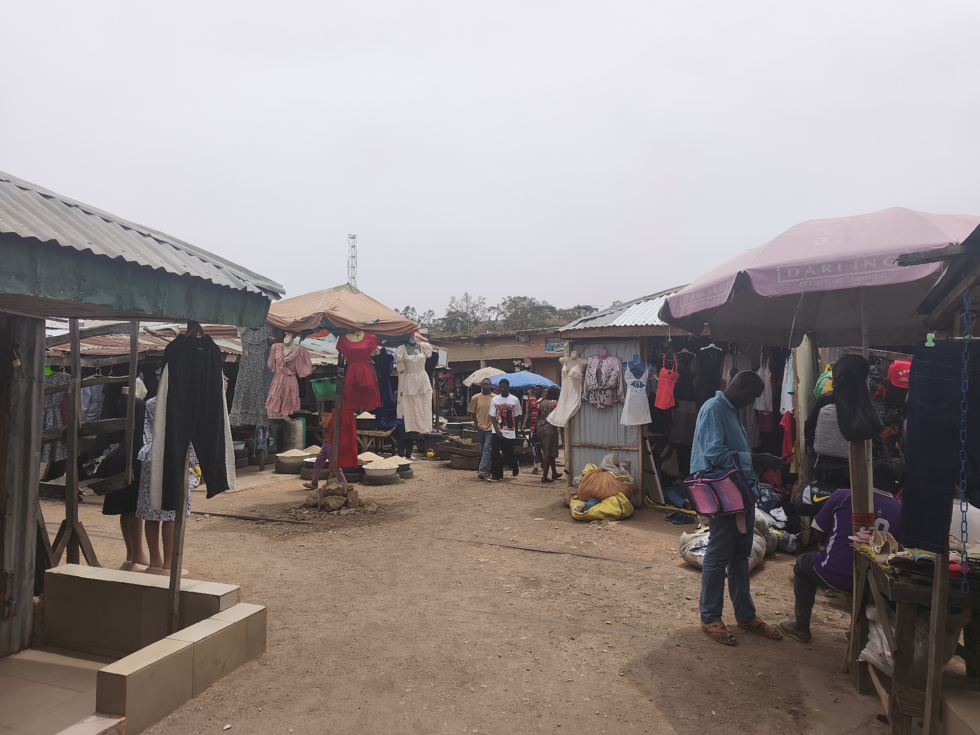 This image captures a bustling outdoor market scene in Jos, Nigeria, under an overcast sky. The ground is unpaved dirt, and numerous market stalls line a central pathway.
On the left side of the frame, several clothing items, including dresses and trousers, hang from wooden structures, with mannequins displaying some garments. The stalls feature corrugated metal roofs, indicative of temporary or semi-permanent structures. Further into the left-center, more clothing is visible, and sacks of what appear to be grains or other goods are arranged on low tables.
Towards the right, a large pink market umbrella provides shade over another stall. Clearly visible on the umbrella are the words "DARLING" and the hashtag "#Find Your Beautiful". Under this umbrella and around the right-side stalls, various pieces of clothing, including dresses, tops, and bags, are displayed. A man in a blue shirt and jeans stands, holding a pink shoulder bag, while another individual is seated nearby.
In the middle ground, several people can be seen walking through the market, engaged in shopping or moving between stalls. The background reveals more market structures, some with corrugated roofs, and distant trees, with a tall communication tower visible against the light grey sky. The overall atmosphere is that of a typical, active market day.