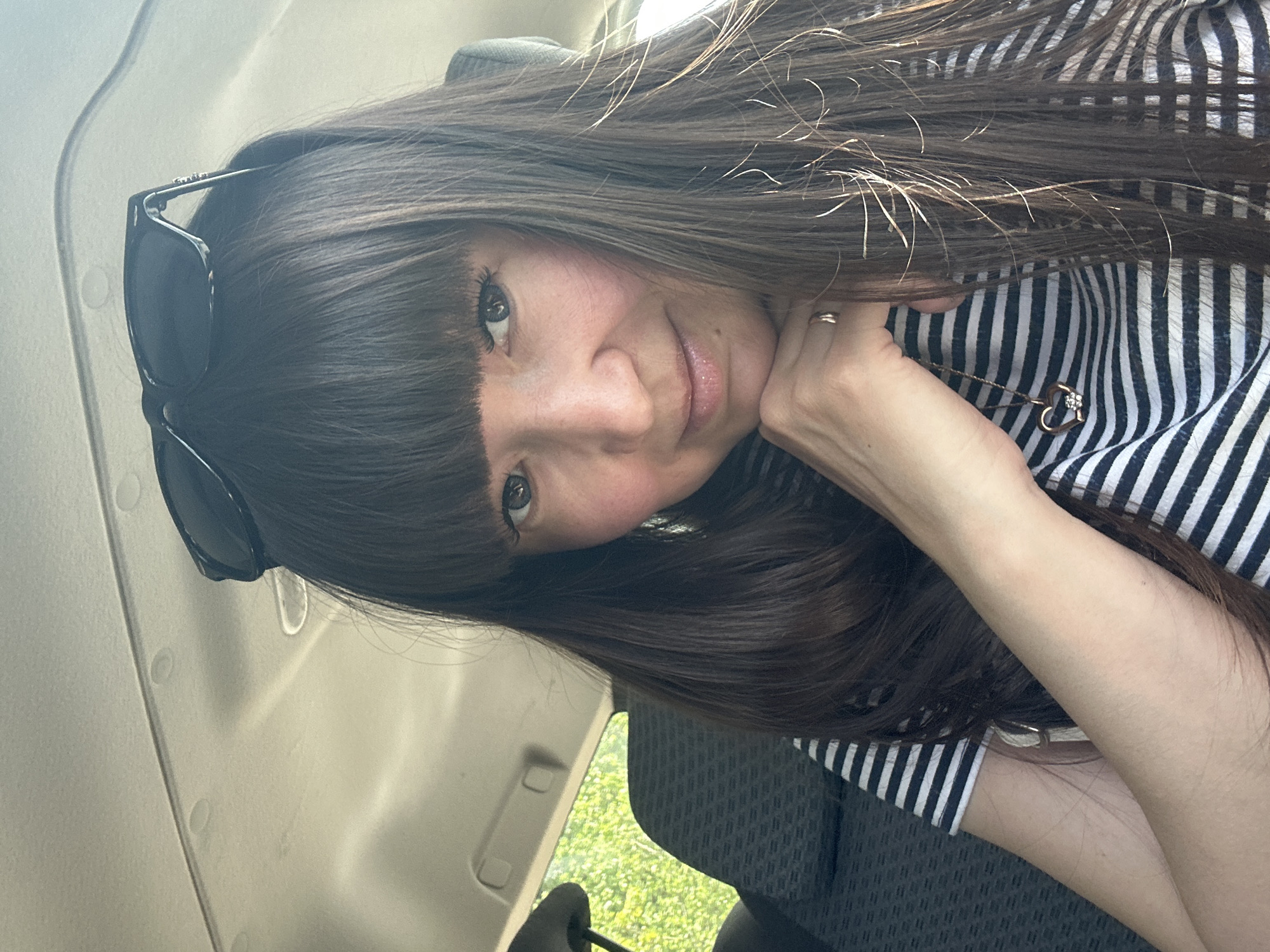 A woman with long, dark hair and bangs is sitting in a vehicle. She is wearing a striped black and white shirt and a gold necklace with a heart pendant. Sunglasses are perched on her head. Her left hand is resting on her chin. Outside the vehicle window is greenery, possibly indicating a park or residential area. The image was taken in a car, which can be inferred the location is Chaikovsky, Russia.