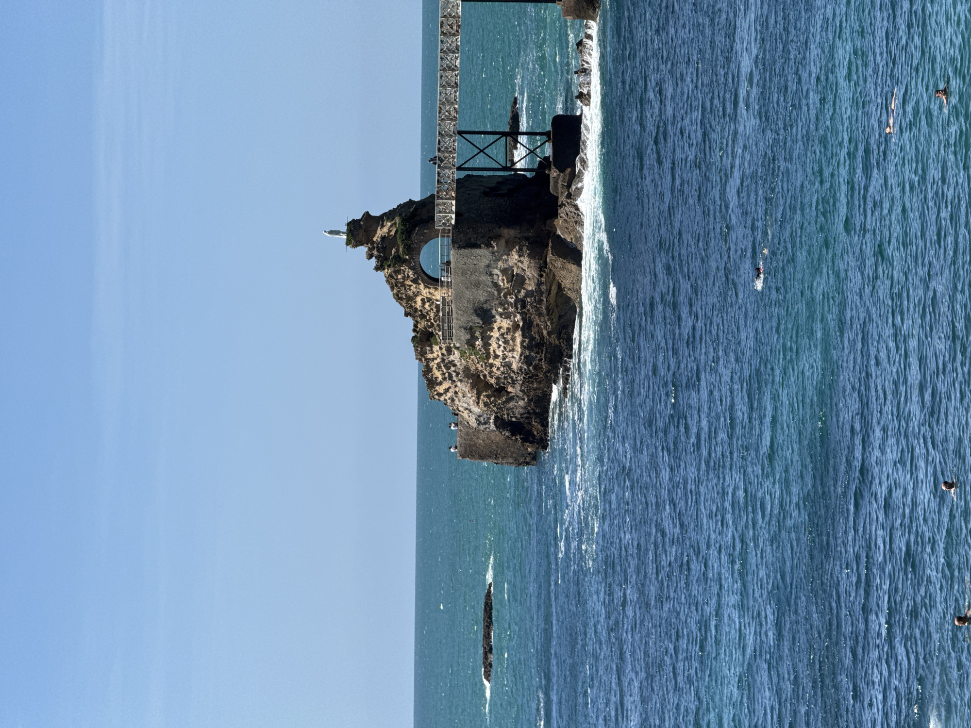 The image captures a sunny day at the Rocher de la Vierge (Rock of the Virgin) in Biarritz, France, a famous landmark situated on a rocky outcrop in the Atlantic Ocean. A narrow, ornate white footbridge, supported by dark metal beams, connects the mainland to the rock. Perched atop the highest point of the rock formation is a white statue of the Virgin Mary, a prominent feature of the site. The rough, textured surface of the rock is visible, with some areas appearing to be reinforced with stone structures. The sea is a vibrant blue, with gentle waves lapping at the base of the rocks and creating white foam. In the foreground, several individuals are swimming or floating in the ocean, enjoying the clear weather. The sky above is a bright, cloudless blue, typical of a summer day.