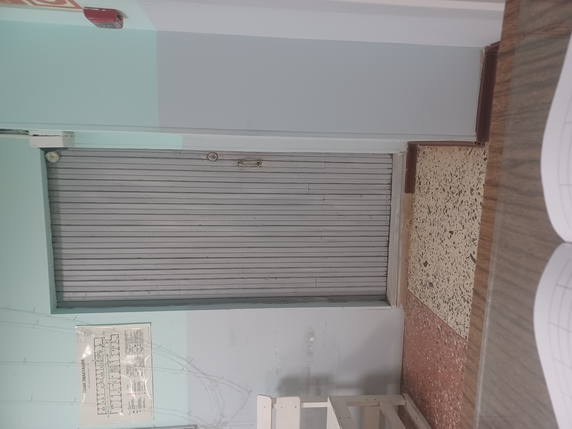 The image depicts an interior hallway with a light blue and grey painted wall, separated horizontally. On the left side of the wall, an evacuation plan is posted. Below the evacuation plan, a white wooden bench is visible. A large, grey metal rolling door dominates the center of the image. To the right of the door, a red fire alarm is mounted on the wall. The floor features a terrazzo pattern, with two distinct sections: one reddish-brown and the other light beige with grey speckles. In the foreground, a wooden surface, possibly a desk or table, is present, with the edge of an open notebook visible. The scene is devoid of people and any discernible activity. The location of Shudayag, Russia, is not visually ascertainable from the image itself.