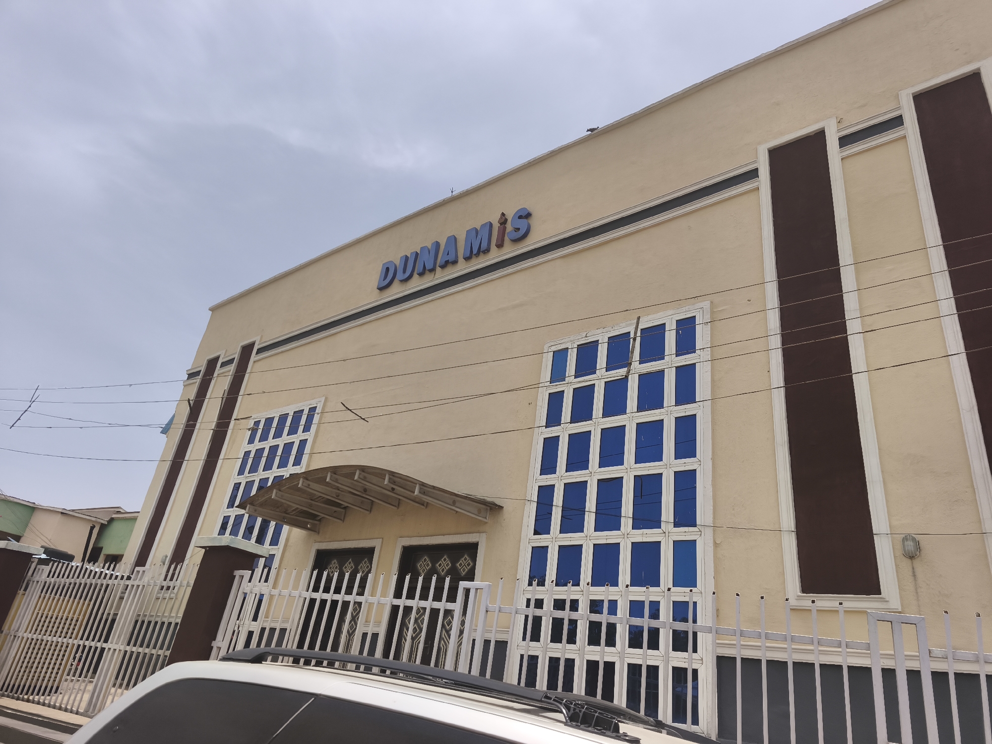 A low-angle, eye-level outdoor shot captures the facade of a building under a bright, overcast sky in Kabong, Nigeria. The building is predominantly light beige or cream in color. Large, blue, three-dimensional letters spelling "DUNAMIS" are prominently affixed to its upper section.
The architectural design includes a thin, dark horizontal stripe running across the facade, just below the name. Vertical brown panels with white borders are integrated into the building's structure, providing a contrasting element. The building features multiple rectangular windows with vibrant blue panes and white frames, arranged in grids, with a larger cluster on the right and a smaller one on the left. A light-colored, curved canopy extends over the main entrance, beneath which two dark, patterned double doors are visible.
In the foreground, a white metal fence with vertical bars and square posts runs across the front of the property. The roof and hood of a white vehicle are partially visible at the bottom of the frame, positioned in front of the fence. The sky is bright and appears cloudy or hazy, indicating daytime. Faint utility lines are present across the upper part of the image. Other blurry structures are visible in the background to the left, suggesting an urban or suburban setting.