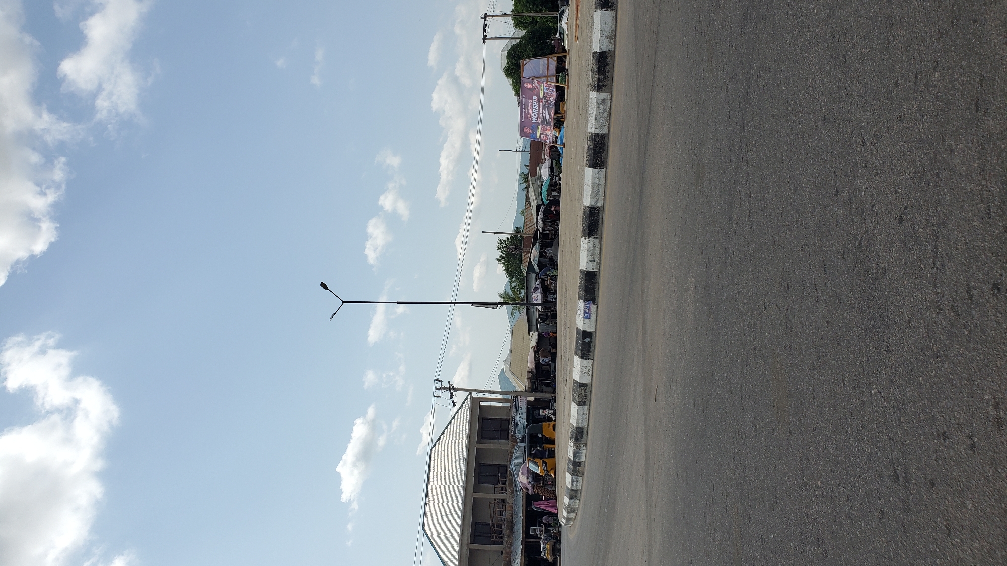 A street scene in Jalingo, Nigeria, features a roundabout with a central grass median bordered by a curb painted with black and white stripes. A tall, black street lamp stands in the center of the roundabout. On the left, a two-story building with a grey, corrugated metal roof and large, dark-tinted windows dominates the foreground. Adjacent to it are market stalls with various umbrellas and awnings, where people are seen browsing. A yellow three-wheeled auto-rickshaw is parked near the building. Further to the right, more market stalls are visible, some with people gathered around them. A large banner with text and images is prominently displayed on the right side of the roundabout, advertising a worship event. The sky is blue with scattered white clouds, and in the background, hills are visible. The road in the foreground is asphalt, showing some wear and tear. The overall scene depicts a busy, outdoor market area in a Nigerian city.