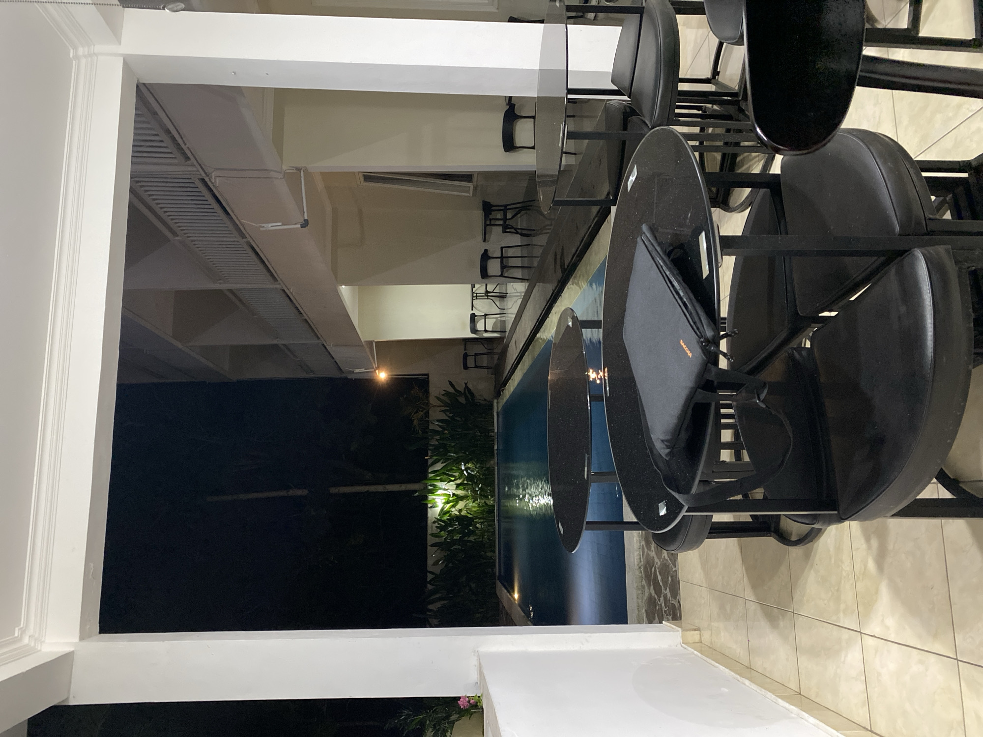 This is a nighttime photograph taken outdoors at a resort in Kuta Selatan, Indonesia, likely a poolside cafe or dining area. In the foreground, there are several black, circular tables with matching chairs. One table has a black folder or portfolio on it. The tables and chairs are made of dark, reflective material. Beyond the seating area is a swimming pool, illuminated from below, with clear blue water. Lush green plants are visible behind the pool, illuminated by spotlights. In the background, the facade of a multi-story building with balconies and railings is visible, also lit by artificial light. The overall atmosphere is serene and tranquil, with the sounds of the night and the gentle illumination of the pool creating a relaxing ambiance.