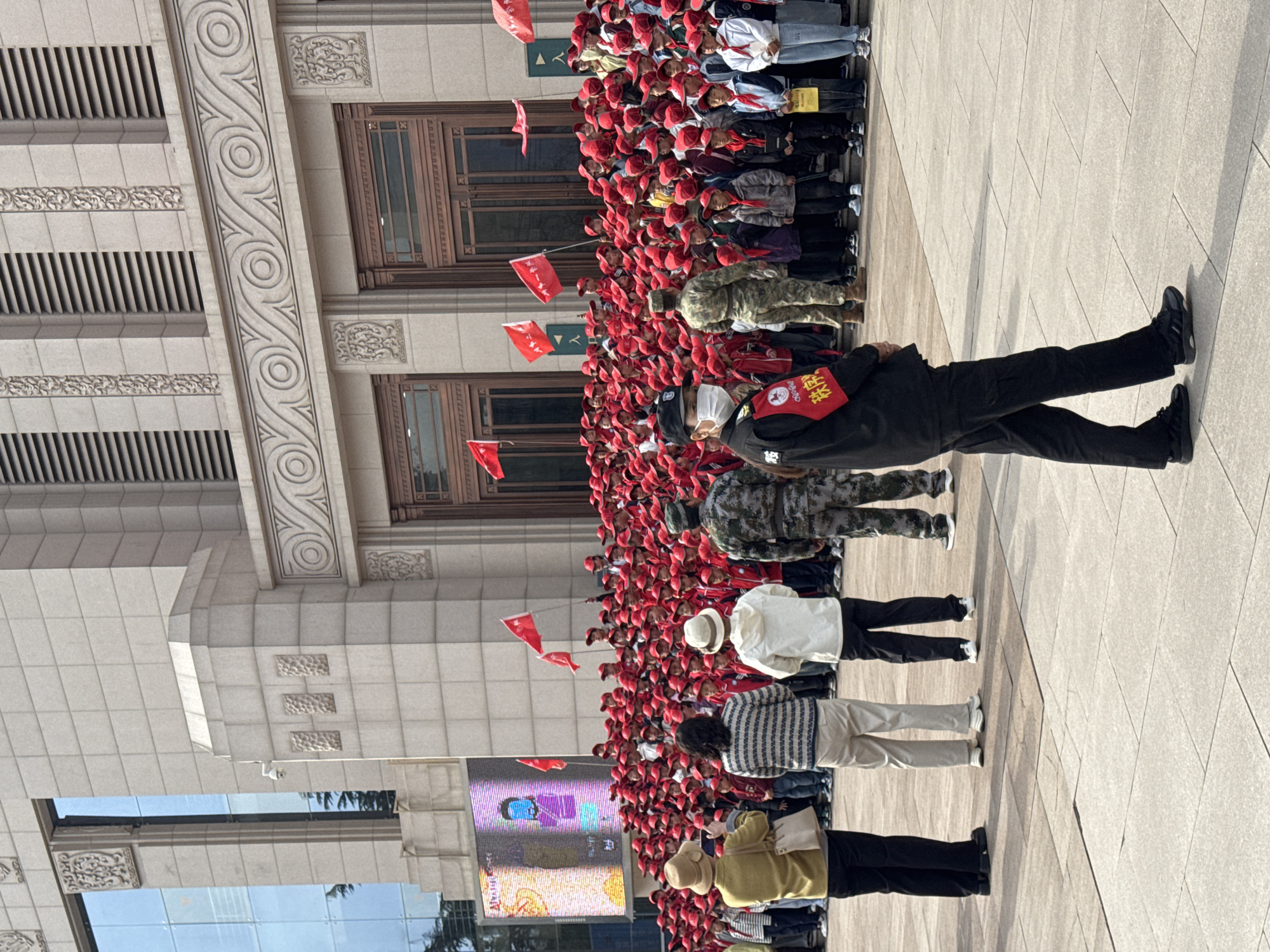 A large group of individuals, mostly children, are standing in front of a building with architectural details. Most children wear red hats and hold small red flags. Adults standing with them include individuals in military camouflage clothing, a person in a black uniform with a red armband, and others in civilian attire. A large digital screen is to the left. The location is identified as Jianlan Road District, China.