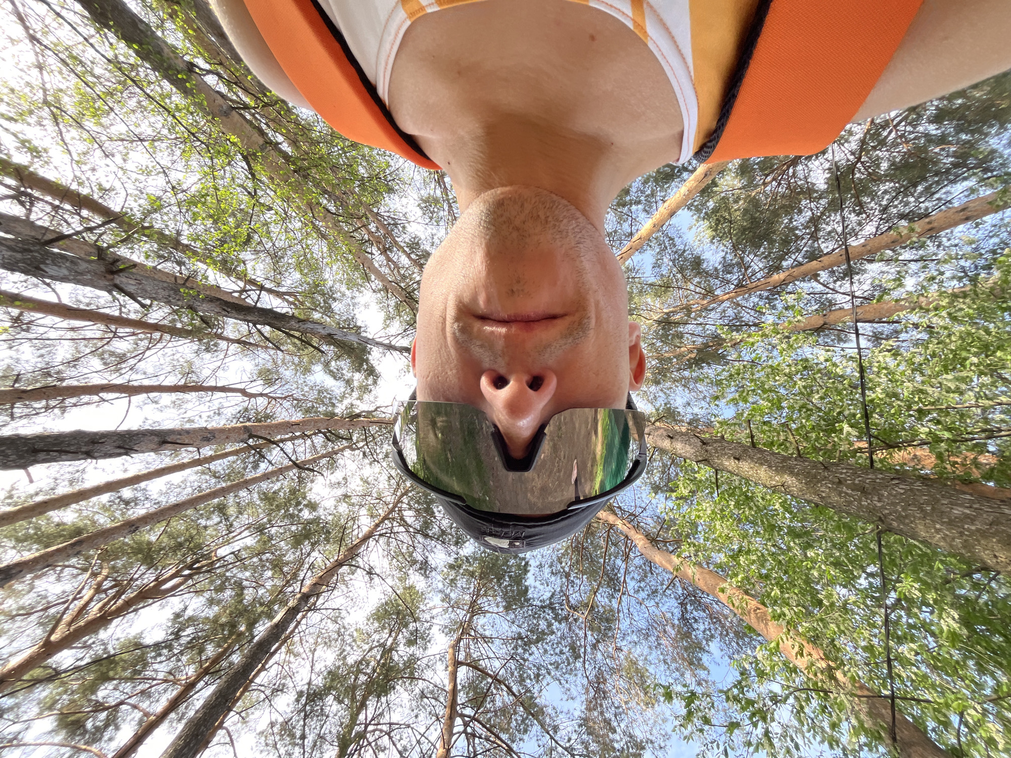 A low-angle, distorted, close-up photograph shows a man with short hair, wearing reflective sunglasses and a light-colored shirt with orange straps, lying on the ground looking upwards. The man's face is the central focus of the image. The background reveals a dense forest canopy of tall trees with slender trunks, and sparse foliage, with a patch of bright sky visible in the upper portion of the frame. Given the perspective, the location is likely outdoors in a forested area. The city is not explicitly recognizable in this image, so its mention cannot be verified.