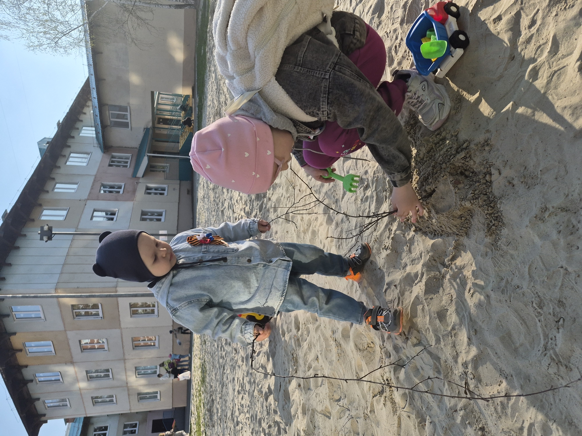Two children are playing in a sand pit located outside a multi-story apartment building. The building features light-colored, possibly pastel, exterior walls with multiple windows.
One child, wearing a pink hat, a light-colored jacket with a grey hooded sweatshirt underneath, and dark pants, is kneeling and playing in the sand with a small green rake. A toy truck and a shoe are also visible in the sand near this child.
The other child, wearing a dark hat, a denim jacket, and jeans, is standing and holding a small object in their hand while also seemingly playing with sticks in the sand.
The setting appears to be in Tobolsk, Russia, as suggested by the location metadata. The weather seems sunny, given the shadows and bright lighting. Other people and activity are vaguely visible in the background near the apartment building.