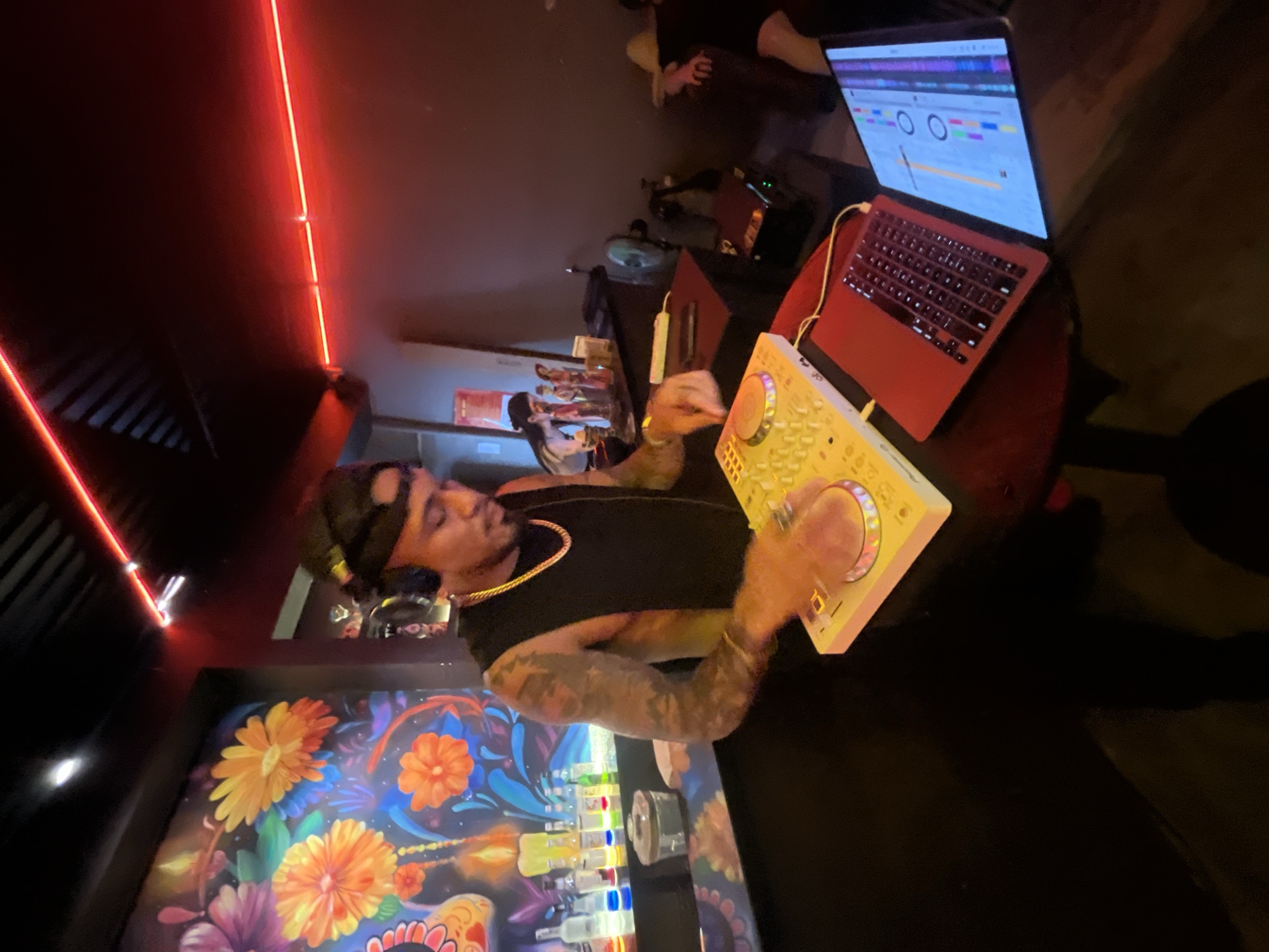 This image depicts a DJ set up in what appears to be a bar or nightclub. A person wearing headphones and a black sleeveless shirt is actively engaged with a white DJ controller, with their hands over the controls. To the right of the DJ controller is a bright red laptop, also open and in use, displaying waveforms and music production software. The background features a vibrant mural with colorful flowers, and a bar area with bottles of liquor and glasses. Red neon lights are prominent, illuminating the space. The overall ambiance suggests a lively evening venue. The location is in the city of Đà Nẵng, Vietnam.