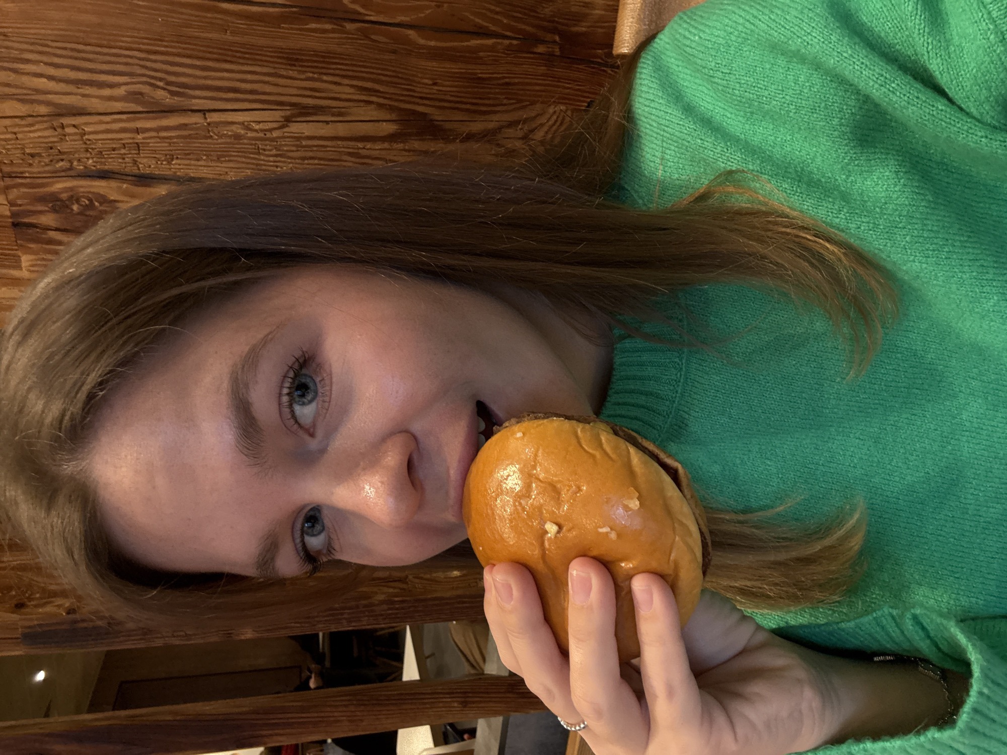 A woman is shown in a close-up shot, tilted to the side, with a hamburger in her hand about to take a bite. She has light skin, blue eyes, and light brown hair. She is wearing a bright green sweater. Her hand, holding the hamburger, has a silver ring on one of the fingers. The background appears to be a wooden surface, possibly a wall or table. The location is not indicated by any landmarks that are typical for Moscow, Russia.