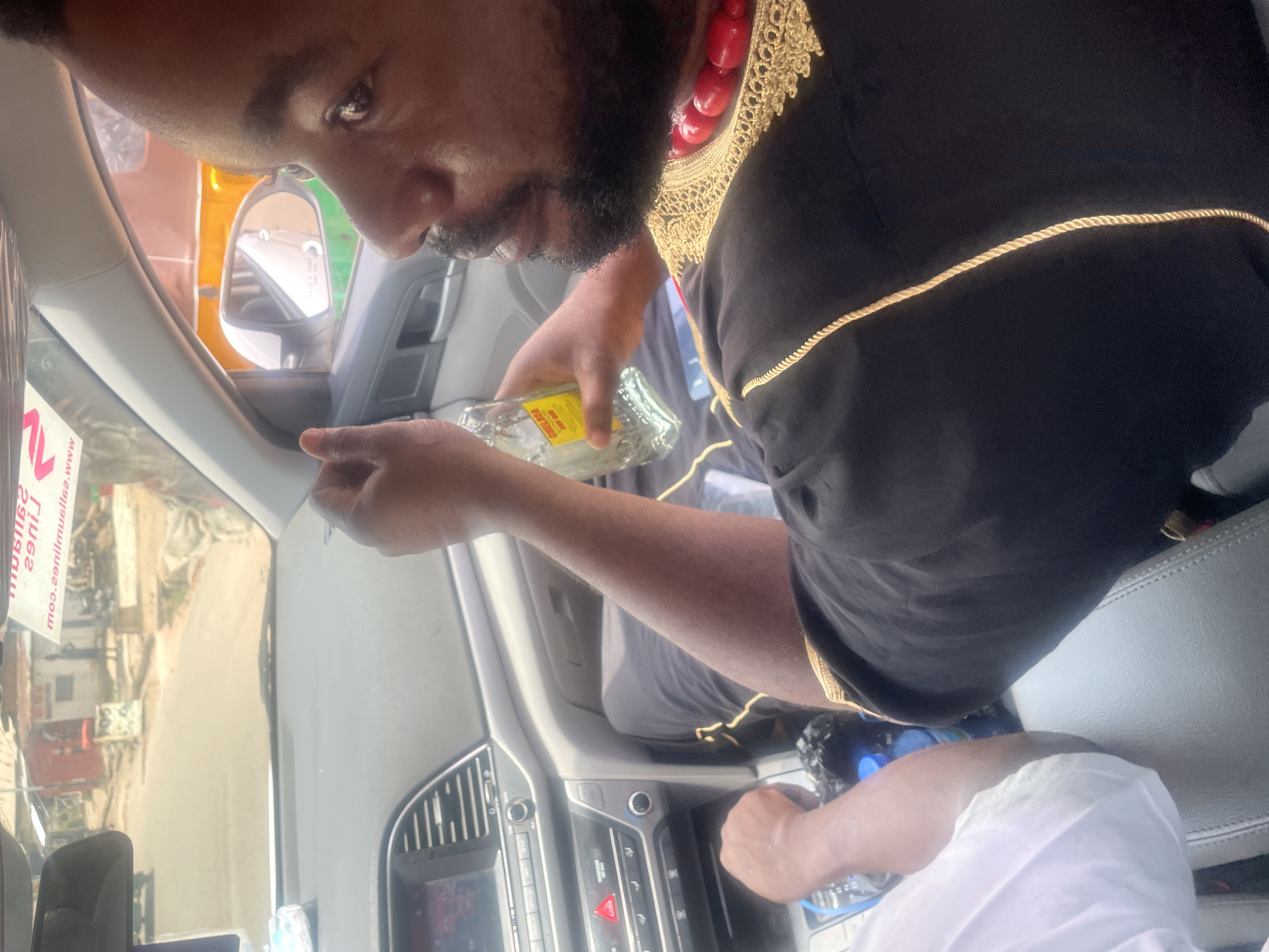 An image shows a man with dark skin and a beard, wearing a black robe with gold trim and a red beaded necklace. He is holding a bottle of what appears to be a yellow liquid, and he seems to be pouring it. He is inside a car, possibly an Hyundai, and the dashboard is visible. Outside the car window, there is a street scene, hinting at an urban area in Jos, Nigeria. The image is rotated, making the scene appear sideways. Another person's arm in a white sleeve is visible in the image.