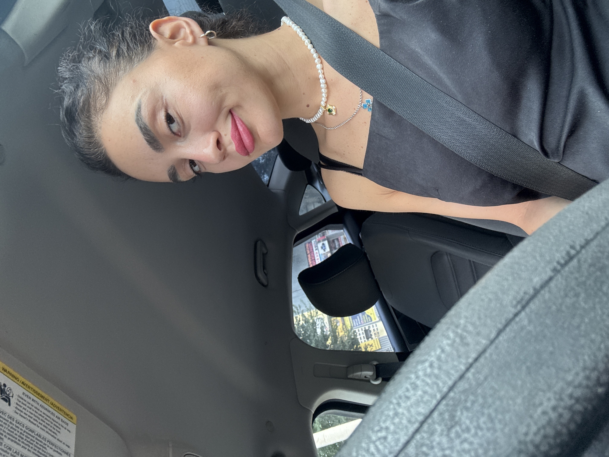 The image is a selfie taken from the driver's seat of a car, likely in Tbilisi, Georgia, given the text visible on a sign in the background which appears to be in Georgian. A young woman with dark hair pulled back and wearing a black dress and a pearl necklace is looking at the camera. She is wearing makeup, including bright pink lipstick. A seatbelt is fastened across her body. The interior of the car is visible, including the steering wheel, dashboard, and passenger seat. Outside the car window, trees and buildings can be seen, suggesting an urban environment. The lighting suggests it is daytime.