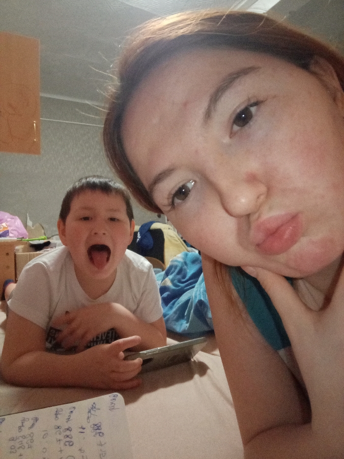 An indoor room in Старый Шагирт, Russia, displays two individuals in a selfie photograph. In the foreground, occupying the right side of the frame, is an adult female with brown hair and dark eyes. She is positioned close to the camera, with her face partially cropped, and is making a "pouty" or "kiss" face with her lips. Her left hand is visible, lightly touching her chin or cheek. She wears a teal-colored top. In the left-mid background, a younger male, approximately a pre-teen or early teen, is lying prone or reclining. He has dark hair and is wearing a white short-sleeved t-shirt. His mouth is open, with his tongue extended. He holds a dark-colored tablet horizontally with both hands. In the immediate foreground, below the male, a white piece of paper with handwritten text and mathematical calculations is visible on a light-colored surface. The background of the room features light-colored, possibly patterned wallpaper. A light brown wooden cabinet is partially visible in the upper left. A blue blanket and other miscellaneous items are present in the mid-background, indicating a domestic setting such as a bedroom or living area. The lighting appears dim.