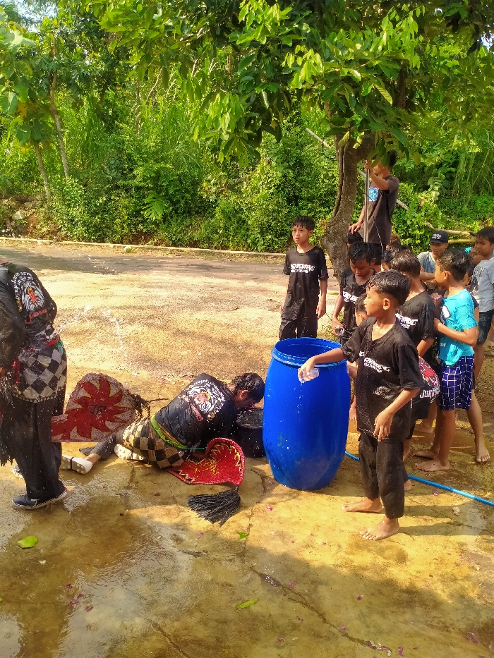 An outdoor scene in Gebyog, Indonesia, depicts approximately a dozen young males engaged in a water-related activity under bright daylight. The foreground features wet, light-colored ground with scattered green leaves and pink petals, indicative of recent water application.
In the left foreground, a person, partially visible, wears dark clothing with a white and black checkered pattern and dark shoes, with water actively splashing off them. Adjacent to this individual, in the mid-ground, a male lies prone on the wet ground, facing right, with his head near a dark bucket. He wears a black t-shirt with a white graphic (possibly an animal head) on the back and checkered trousers. Water splashes around him. Next to him on the ground are two red, fan-like objects with intricate patterns, and a smaller black object with tassels, suggesting performance attire or props.
To the right, a large blue plastic barrel stands upright. A young boy, barefoot and dressed in a dark t-shirt and dark pants, reaches into the barrel with a small white object, water dripping from his hand. Behind him and extending to the right, a group of approximately 7-8 other young boys and teenagers observe the activity. Most are dressed in dark t-shirts and shorts; some are barefoot. One boy in a light blue t-shirt and plaid shorts is prominent in this group. Another boy is seen leaning against or partially obscured by a tree trunk in the upper right. Further back, another boy in a dark t-shirt stands observing.
The background consists of dense green foliage, with larger trees providing shade overhead. A dirt or unpaved road is visible behind the group. A blue hose or pipe lies on the wet ground in the lower right quadrant. The scene suggests a communal activity involving water, potentially a cleansing ritual or post-performance dousing.