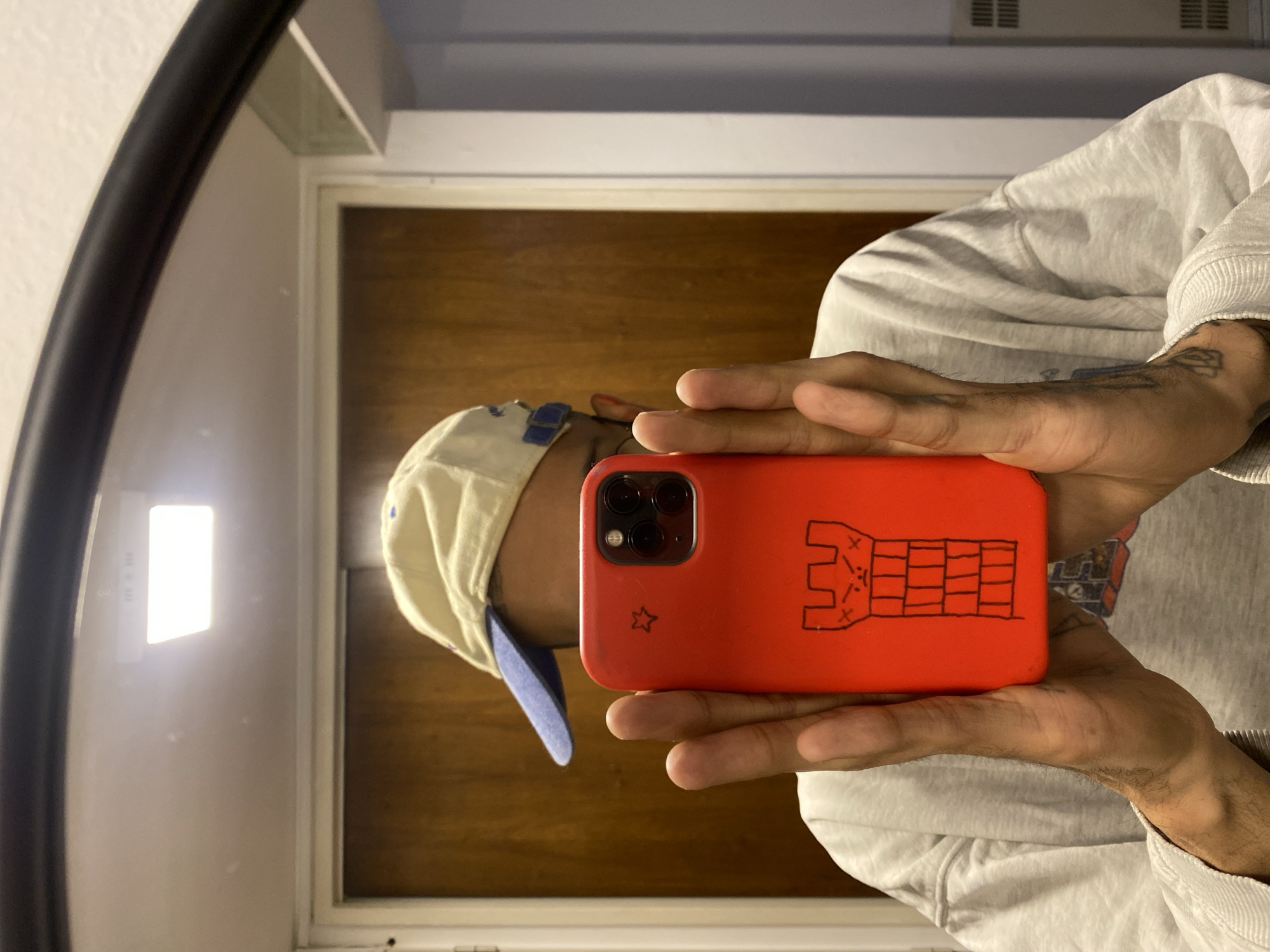 This image is a mirror selfie, rotated 90 degrees counter-clockwise. It shows a person from the chest up, reflected in a large, black-framed mirror, holding a smartphone to capture their reflection.
The subject is wearing a light-colored baseball cap with a blue brim, and a light grey hooded sweatshirt. Their hands, which are holding the phone, display visible tattoos on the back of the hands and forearms. The person's face is mostly obscured by the phone, but part of their profile with glasses is visible behind it.
The smartphone is encased in a bright red protective cover. On the back of the red case, there is a distinct, hand-drawn design in black marker: a blocky, castle-like structure or tower with crenellations at the top and a brick pattern, along with a small star drawn near the bottom left corner of the case. The phone's multi-lens camera array is also visible on the top left of the case.
In the background reflection, a light brown wooden door with a white frame is visible. Above and to the left of the door, a bright, rectangular light fixture is mounted on the wall. Faint, illegible text, possibly "LED 312" or "LED 318," can be seen on this fixture. The overall setting appears to be an indoor space, likely a room or hallway. The location context for the image is Buenos Aires, Argentina. No specific time of day or weather conditions can be discerned from the indoor scene.