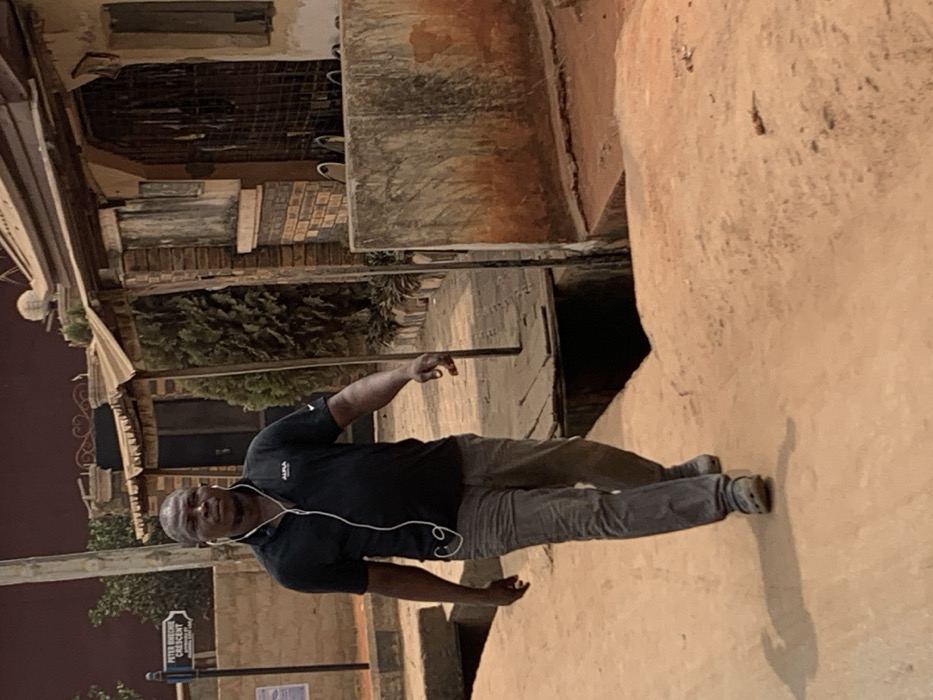 An adult male is captured walking on a reddish-brown dirt path in Otuocha, Nigeria. He is centrally positioned, moving forward and slightly to the right. The man wears a black short-sleeved polo shirt with a white logo on the left chest, grey trousers, and light-colored closed-toe shoes. White wired earphones are visible, with the wires extending from his ears down his torso. To the left, a street sign is visible, identifying "PETER OKECHE CRESCENT" and "MADONA SAFE LANE." An open concrete drainage ditch runs alongside the path on the right, partially bridged by wooden planks. Beyond the ditch to the right, a structure with a corrugated metal roof, exposed brickwork, and a barred opening is present. Green foliage and additional building structures are visible in the background. The scene is illuminated by daylight.