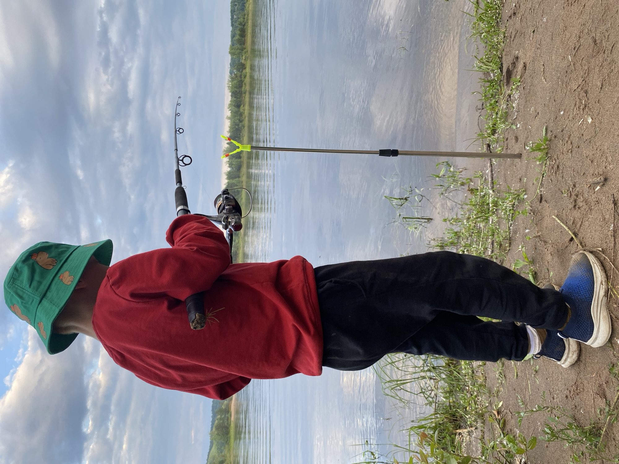 A person, seen from the side, stands on a sandy riverbank, holding a fishing rod. They are wearing a red hoodie, black pants, blue and white sneakers, and a green bucket hat with a cartoonish duck pattern. The fishing rod is silver with black accents and has a reel attached. A yellow fishing rod holder is set up on the bank. The scene appears to be a natural outdoor setting, likely a river or lake, with calm water reflecting a cloudy sky. Trees and distant hills form the backdrop. The overall impression is of a peaceful, recreational activity. The time of day is likely late afternoon or early evening, given the soft lighting and cloudy conditions. There is no visible text in the image.
