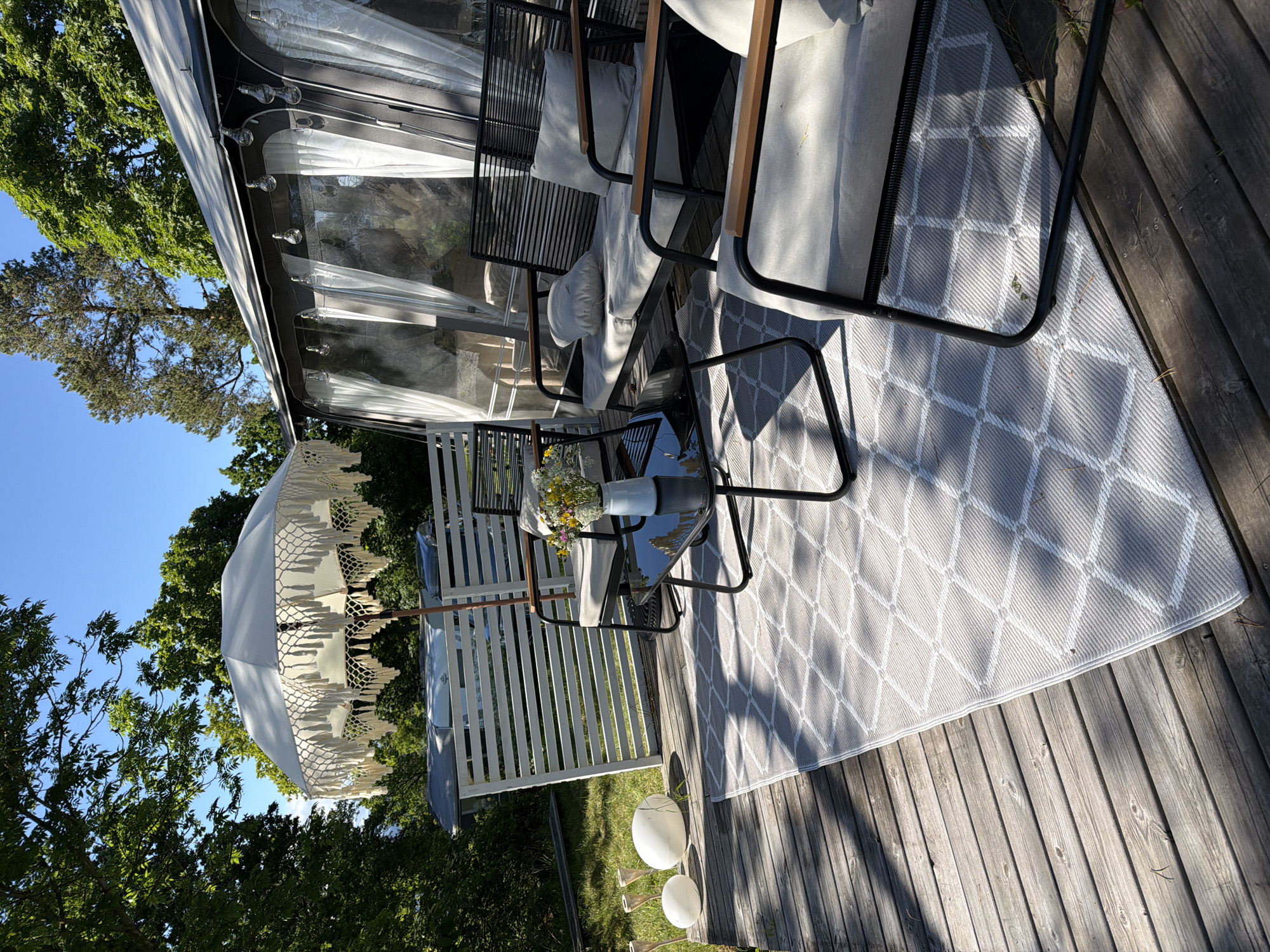 This image captures an outdoor leisure area, specifically a wooden deck, situated in Horten, Norway. The scene is illuminated by direct sunlight, casting distinct shadows across the surfaces.
In the foreground, a weathered wooden deck extends across the bottom and left side of the frame. A rectangular outdoor rug, grey with a repeating white diamond pattern, covers a significant portion of the deck. On the rug, a set of outdoor furniture includes a two-seater sofa and an armchair, both constructed with black metal frames, light grey fabric cushions, and wooden armrest accents. A black, rectangular coffee table with a reflective surface is positioned in front of the sofa. A small, light-colored pitcher containing a bouquet of assorted wildflowers, including yellow, white, and purple blooms, rests on the coffee table.
To the left of the seating arrangement, near the edge of the deck and a white slatted fence, are two large, smooth white spherical objects and several conical, torch-like elements with light-colored tips. The white slatted fence forms a barrier between the deck and a patch of green grass beyond.
Overhead, partially obscuring the view of the sky, is a large white parasol featuring an elaborate macrame fringe and tassels along its edge. Behind the fence and grass, a dark-colored caravan or mobile home is visible, featuring an attached awning structure with transparent plastic walls, internal white sheer curtains, and a string of clear spherical bulb lights suspended along its upper edge.
The background consists of dense green foliage from numerous trees, with glimpses of a clear, vibrant blue sky above. No individuals are present within the frame.