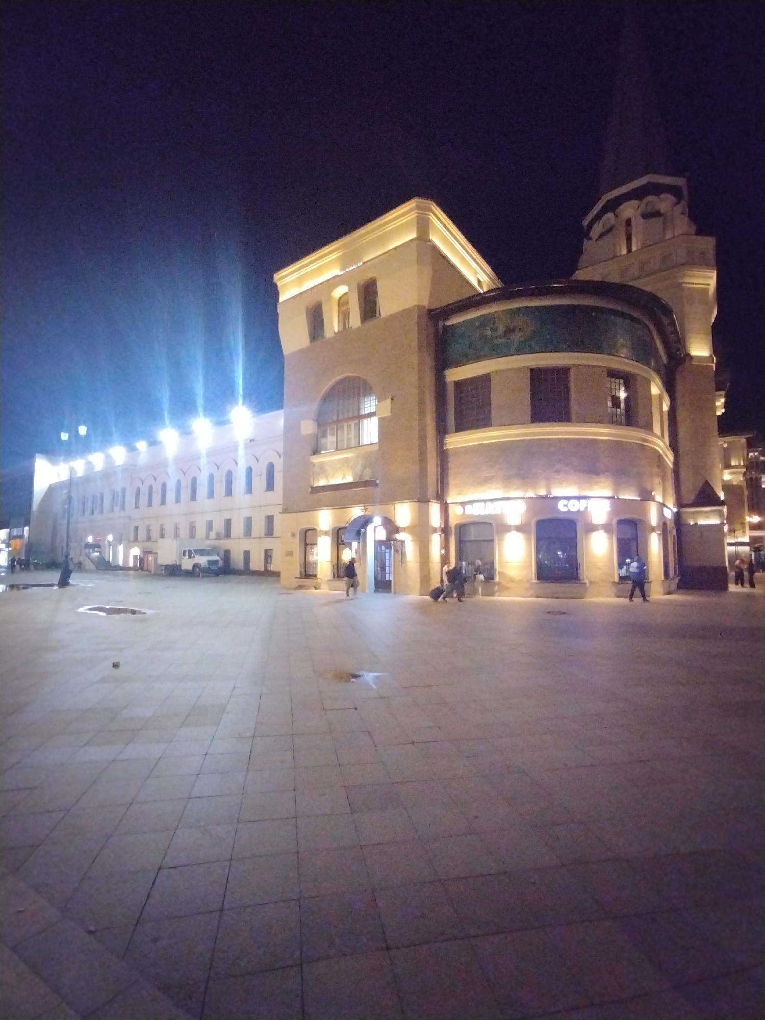 This image depicts a nighttime view of the Kazansky railway station in Moscow, Russia. The station's facade is illuminated, casting a warm glow on the paved plaza in front. The architecture features a long, arched structure on the left and a curved section with a prominent "COFFEE" sign on the right, suggesting a commercial establishment within. Several people are visible, some walking and others sitting or standing near the building, indicating a lively atmosphere despite the late hour. A white vehicle is parked in front of the longer section of the building. The sky above is dark, typical of a nighttime scene.